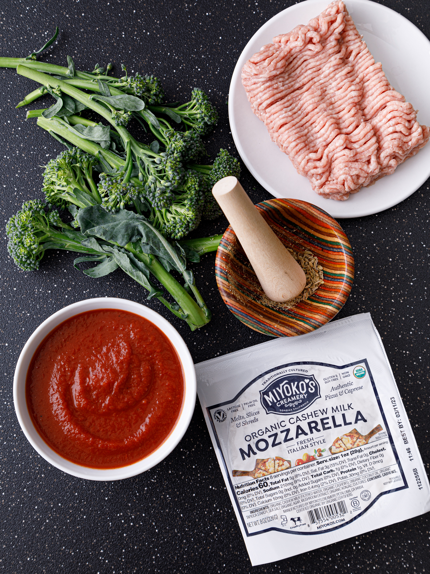 a bowl of tomato free pizza sauce, vegan mozzarella, fresh broccolini. ground turkey and a mortar and pestle with herbs on a dark tabletop