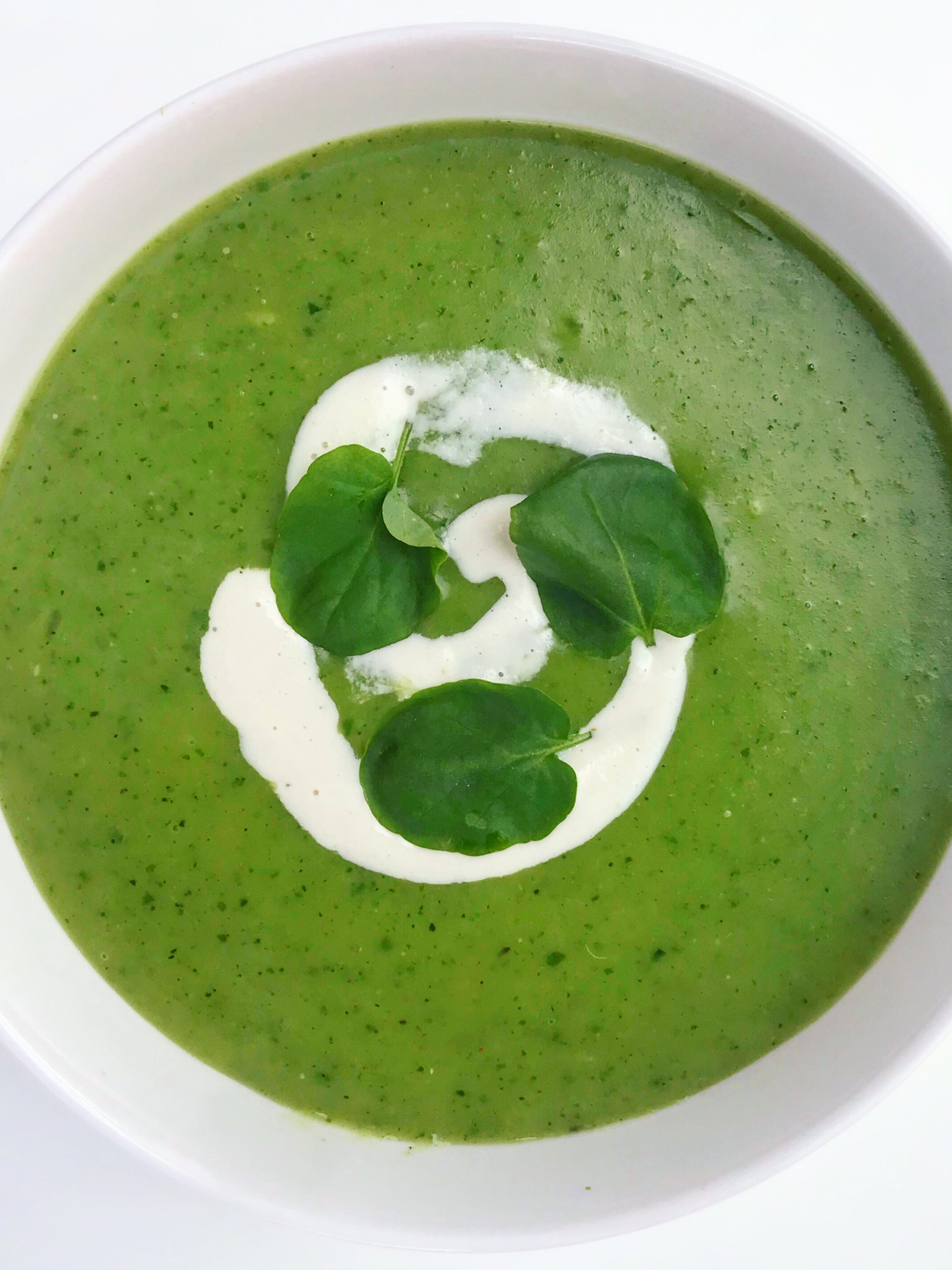 white bowl on a white table full of zucchini and watercress soup with a dollop of cashew cream and topped with fresh herbs