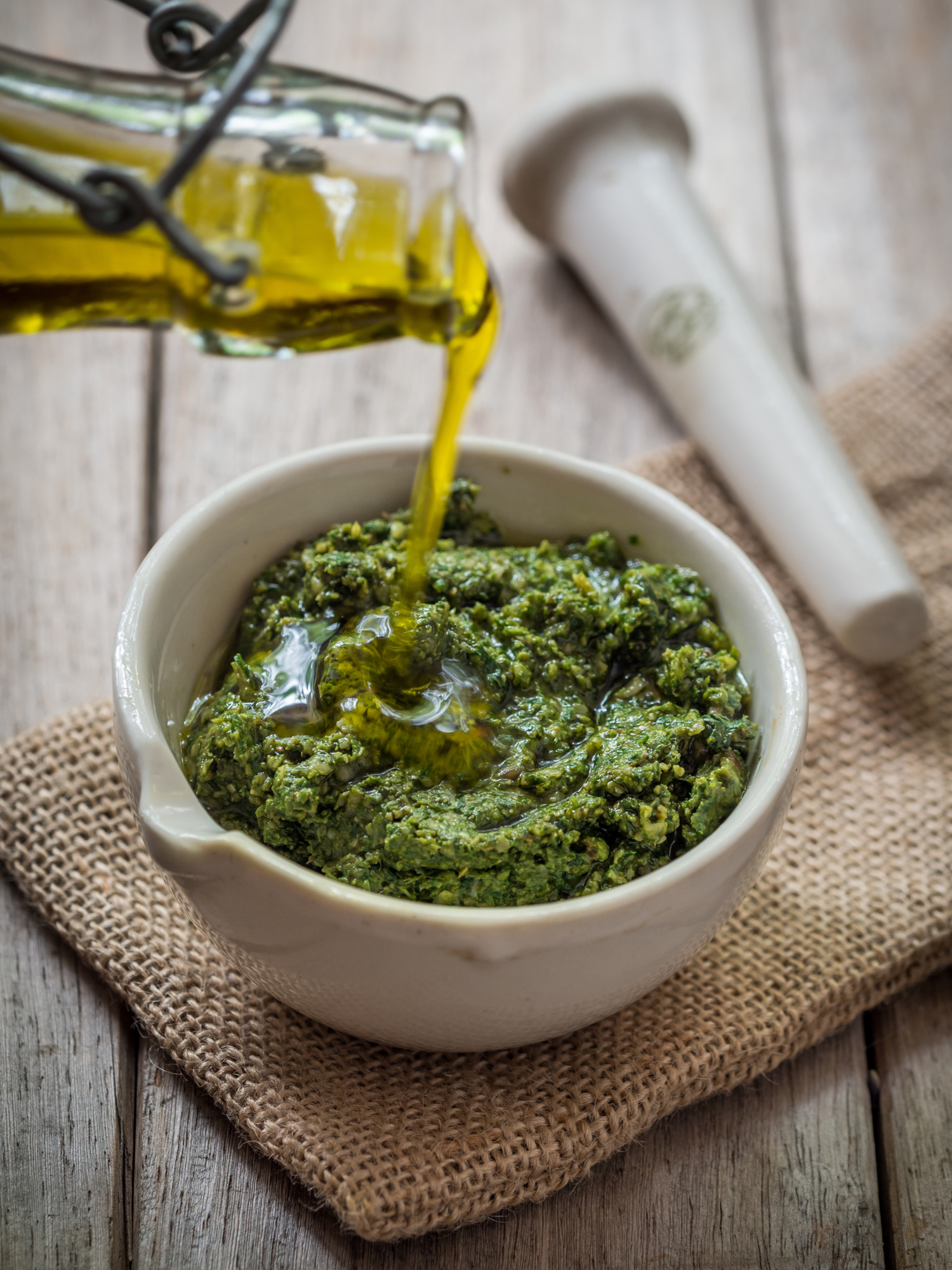 a white bowl full of pesto with olive oil being poured over it sitting on a table with a pestle laying next to the bowl
