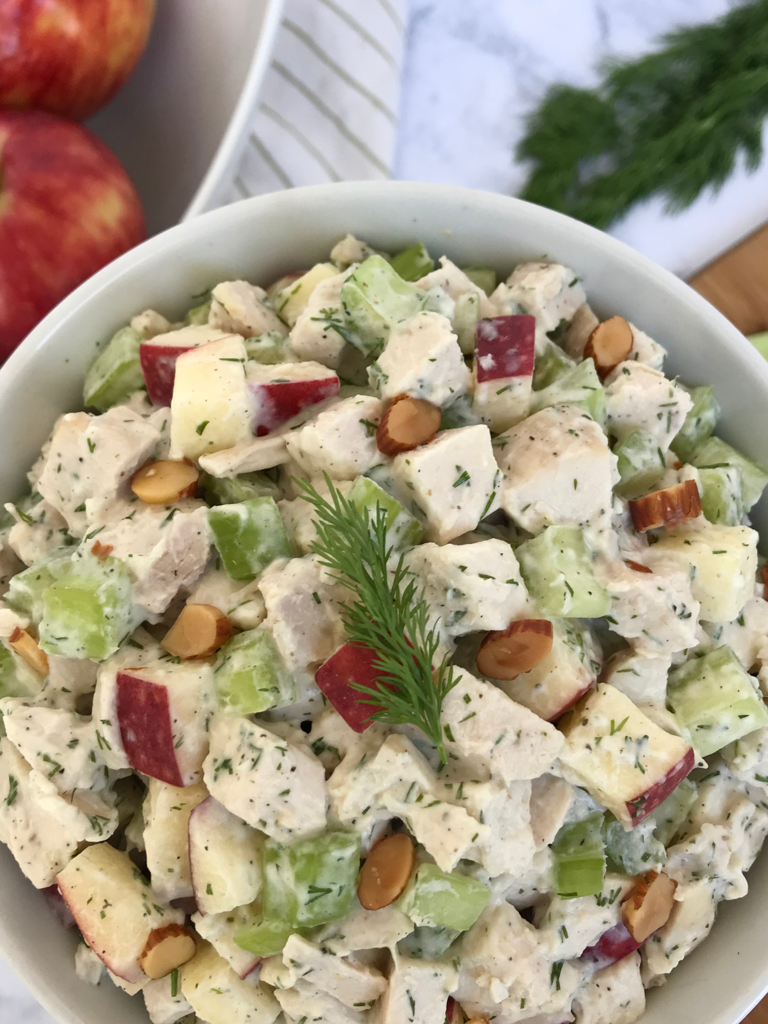 A bowl of Fresh Dill Chicken Salad, with apples, fresh dill, celery, and a cutting board in the background