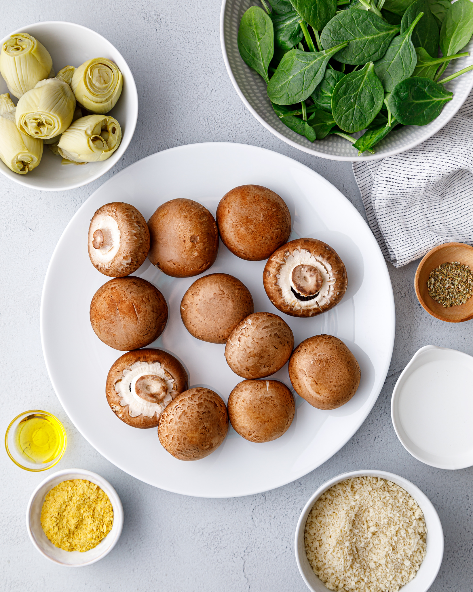 baby bella mushrooms sitting on a white plate surrounded by bowls filled with spinach, artichokes, olive oil and nutritional yeast