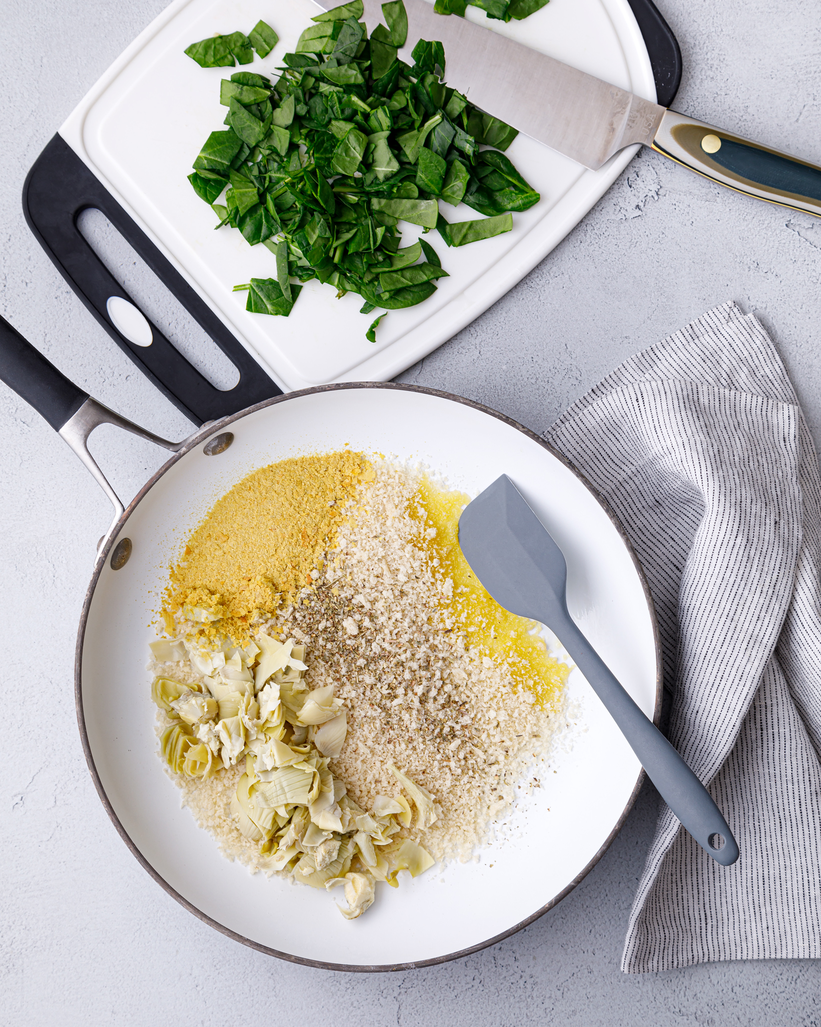 a white bowl with artichokes, gluten free breadcrumbs, olive oil and nutritional yeast with a blue spatula and a cutting board of chopped parsley behind it