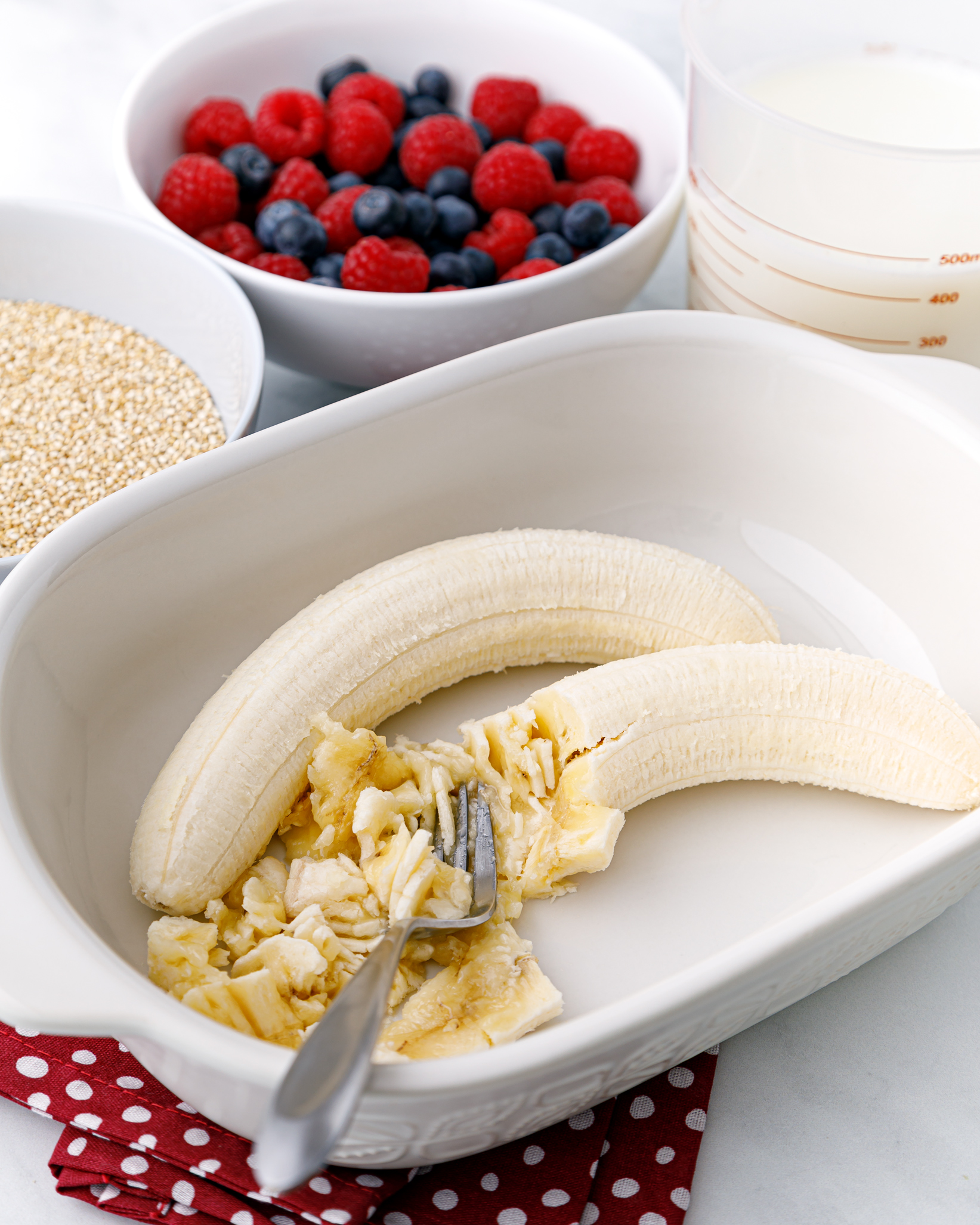 a white casserole dish with two peeled bananas partially mashed and a fork lying in the dish. There is a white bowl with fresh berries as well as a bowl of dry quinoa behind it.