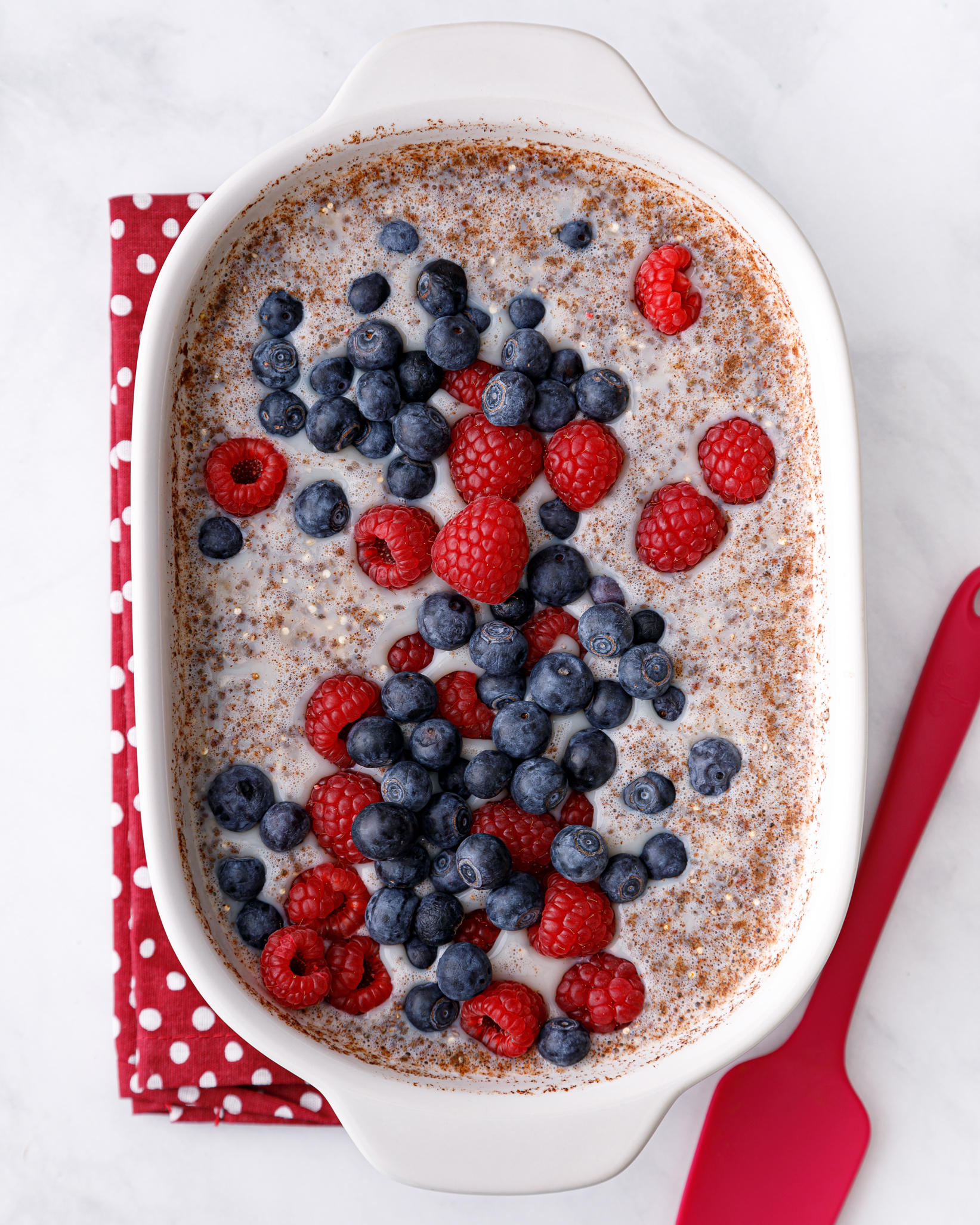 a 2 quart casserole dish with breakfast quinoa bake mixture topped with fresh berries on a red and white polka dot napkin with a red spatula