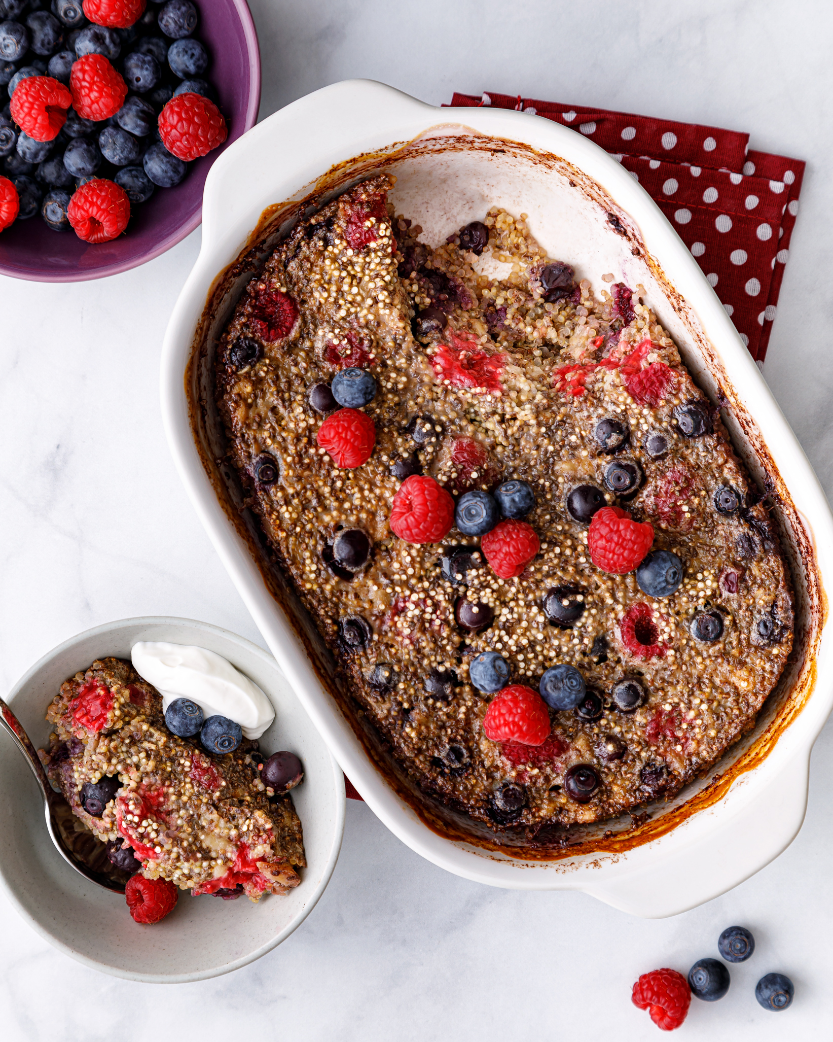 a white casserole dish of quinoa breakfst bake topped with fresh blueberries and rasberries next to a bowl of fresh blueberries and raspberries