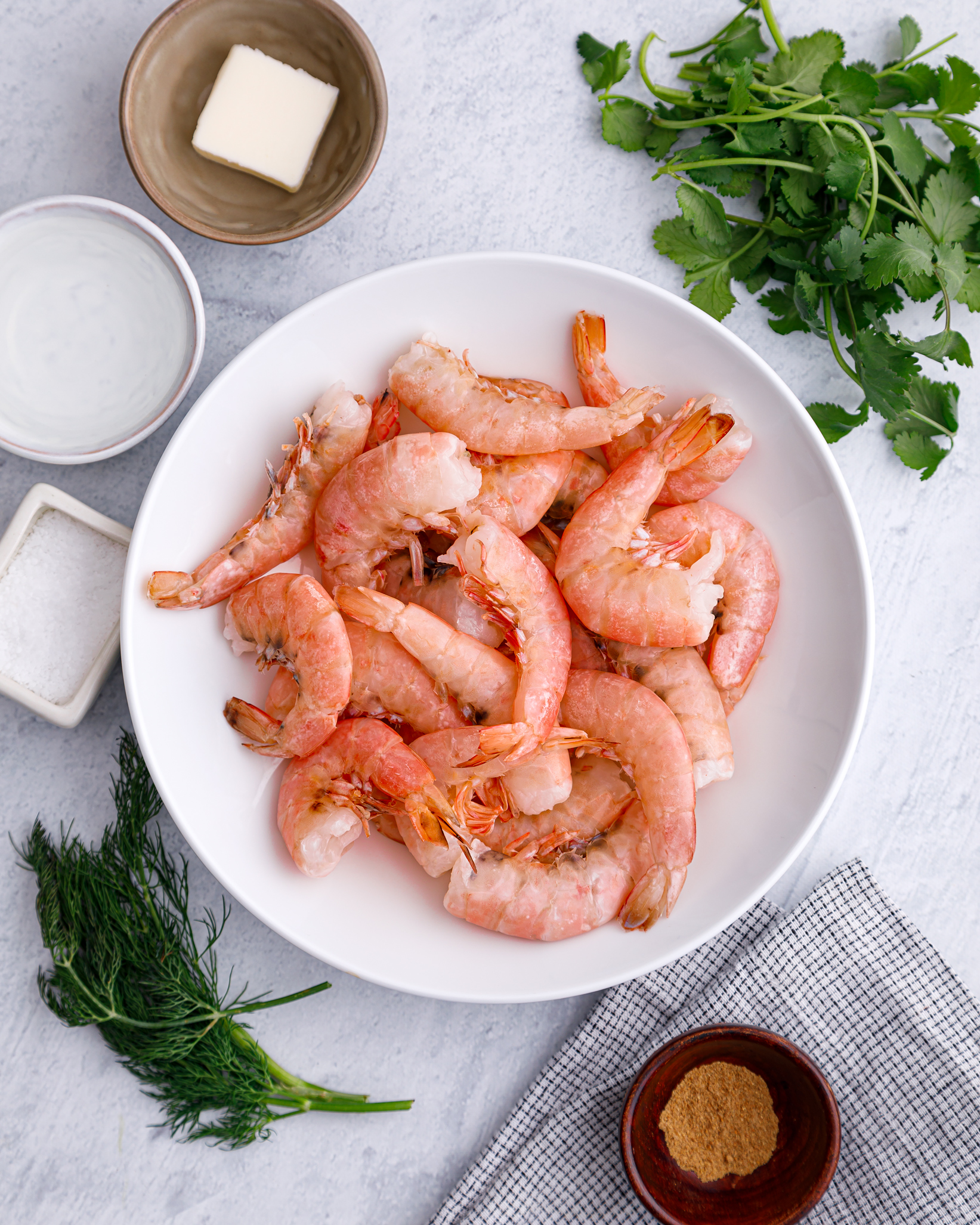 raw argentine red shrimp in a white bowl surrounded by fresh herbs, vegan butter, coriander, salt and other seasosnings