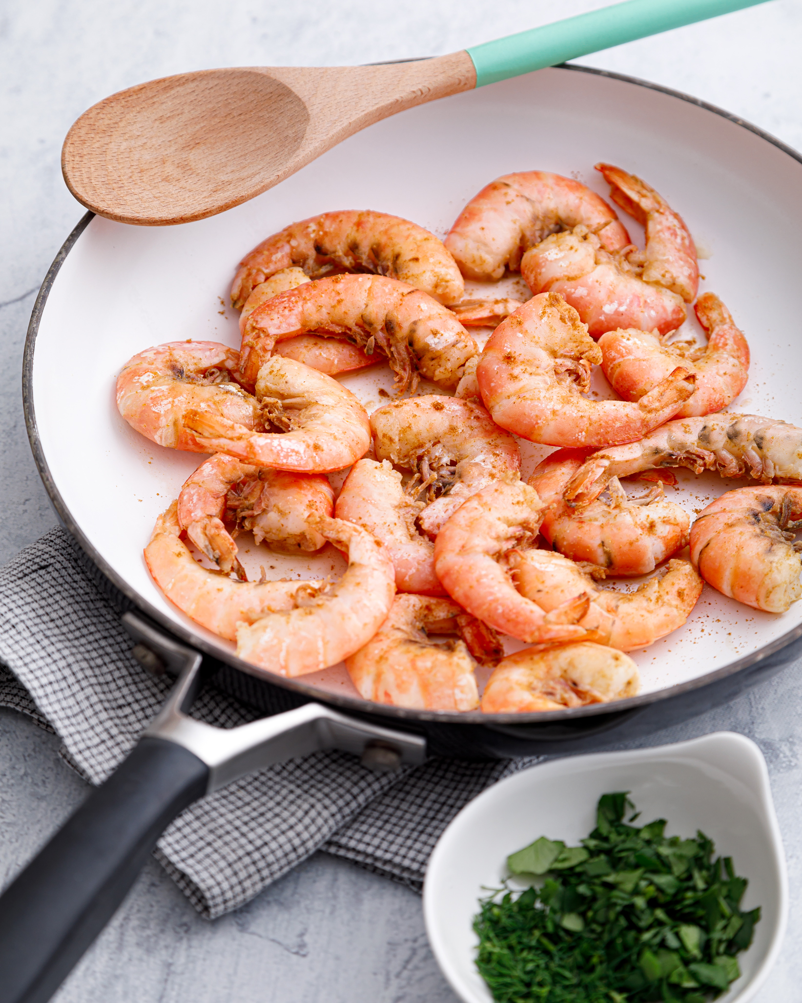 a white skillet of just cooked Argentine red shrimp sitting on lightly colored dish cloths with a wooden spoon and fresh cilantro just barely visible in the picture
