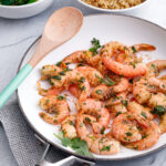 a white skillet of cooked argentine red shrimp sprinkled with fresh herbs, with a wooden spoon laying on top and side dishes in the background