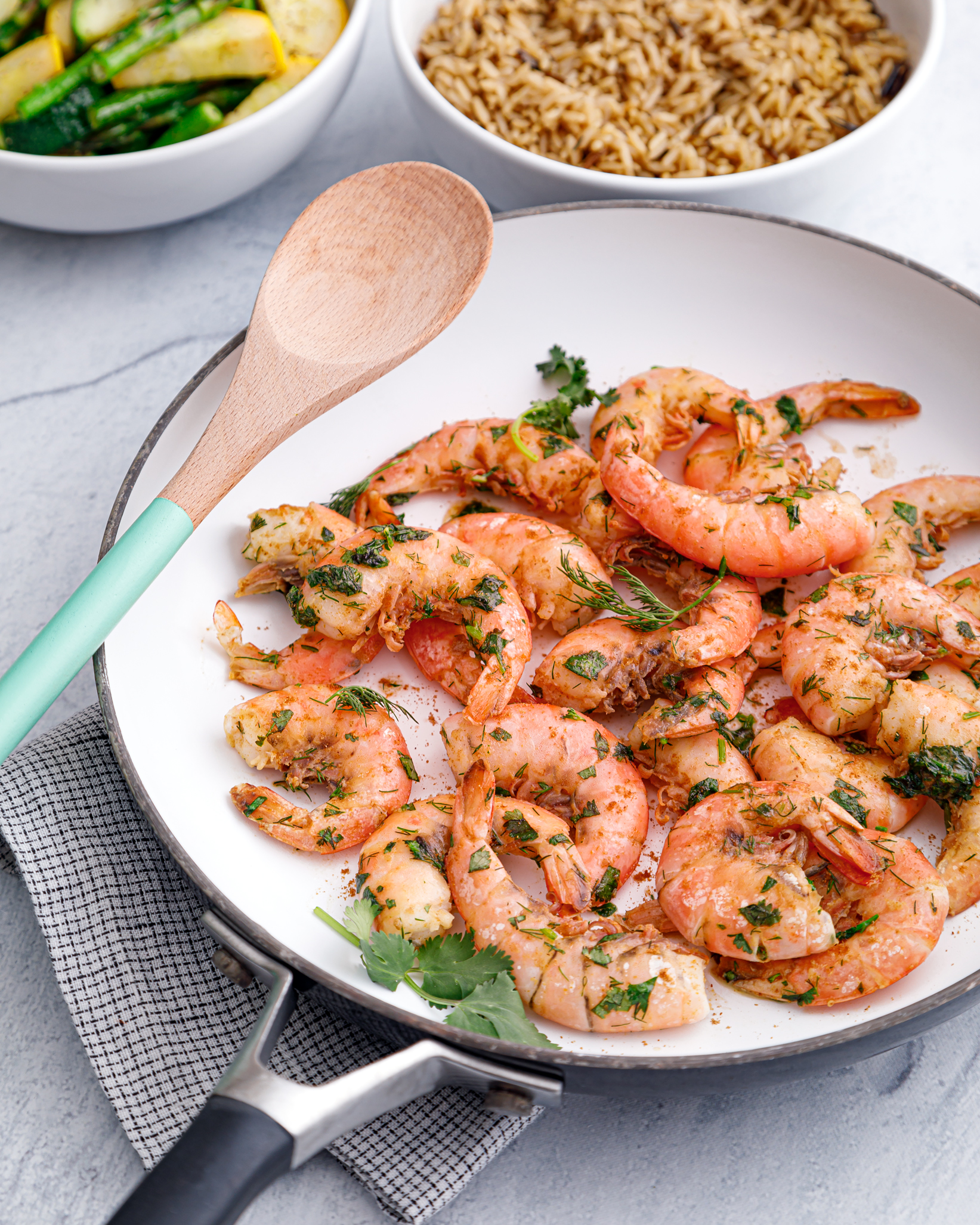 a white skillet of cooked argentine red shrimp sprinkled with fresh herbs, with a wooden spoon laying on top and side dishes in the background