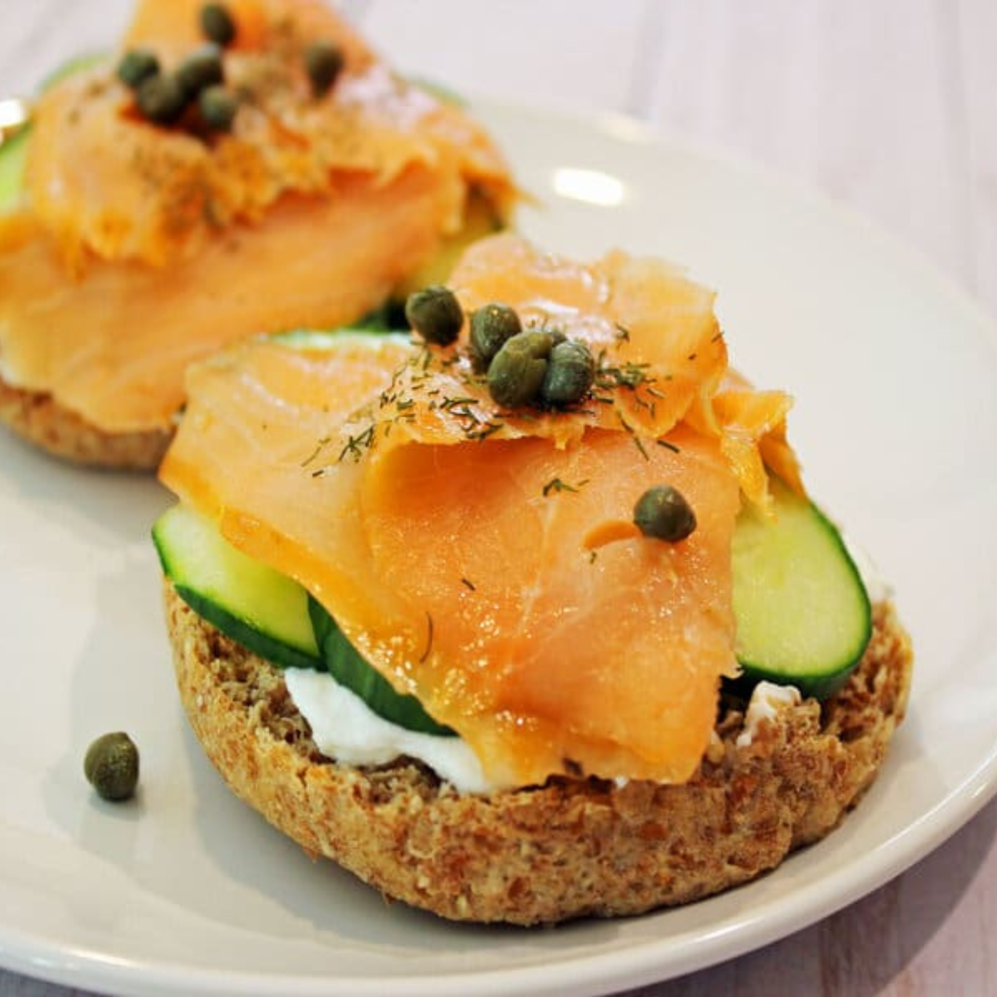 two english muffins topped with greek yogurt, cucumber, smoked salmon, capers and dill on a white plate