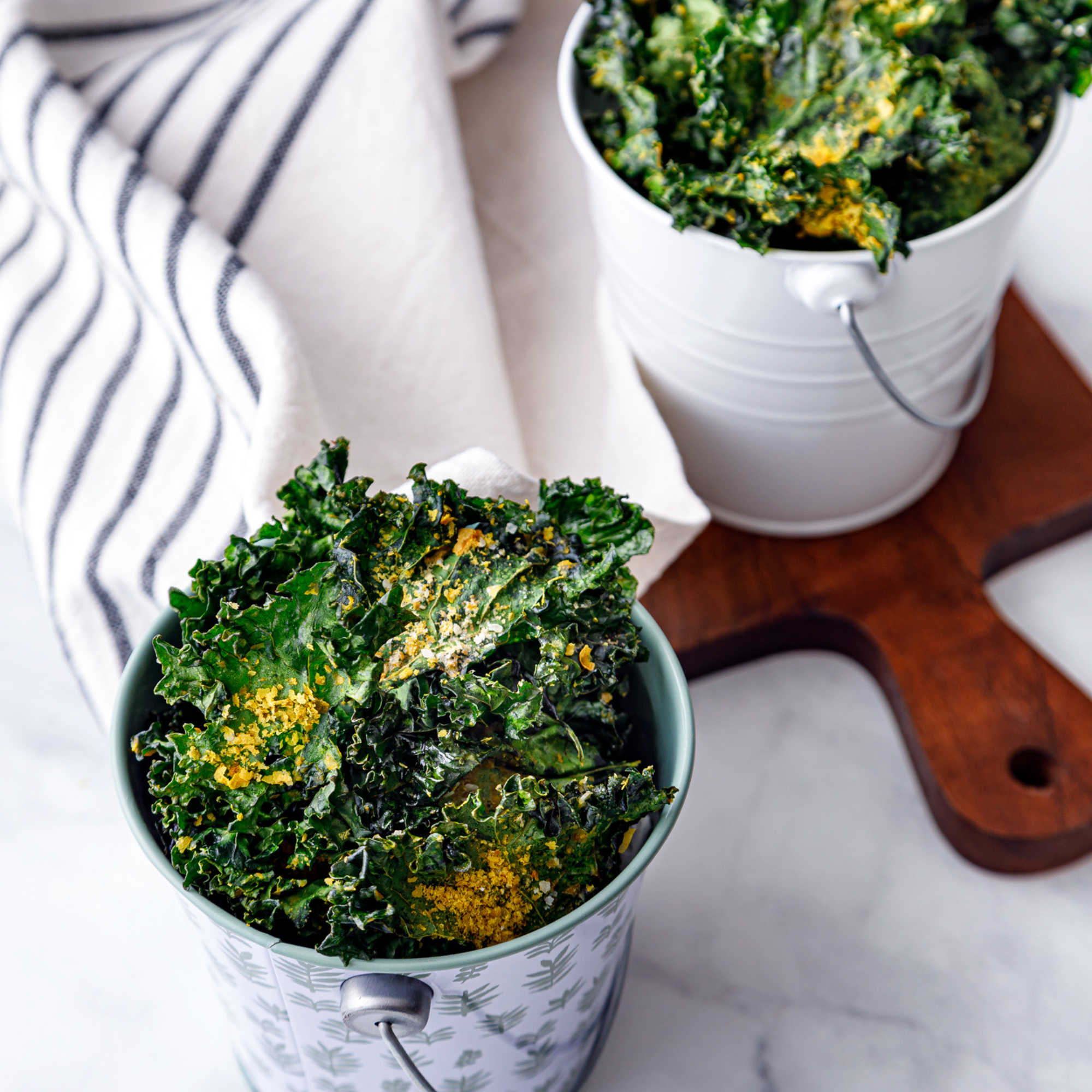 two buckets of kale chips sprinkled with nutritional yeast sitting on a white table top with a blue and white striped cloth