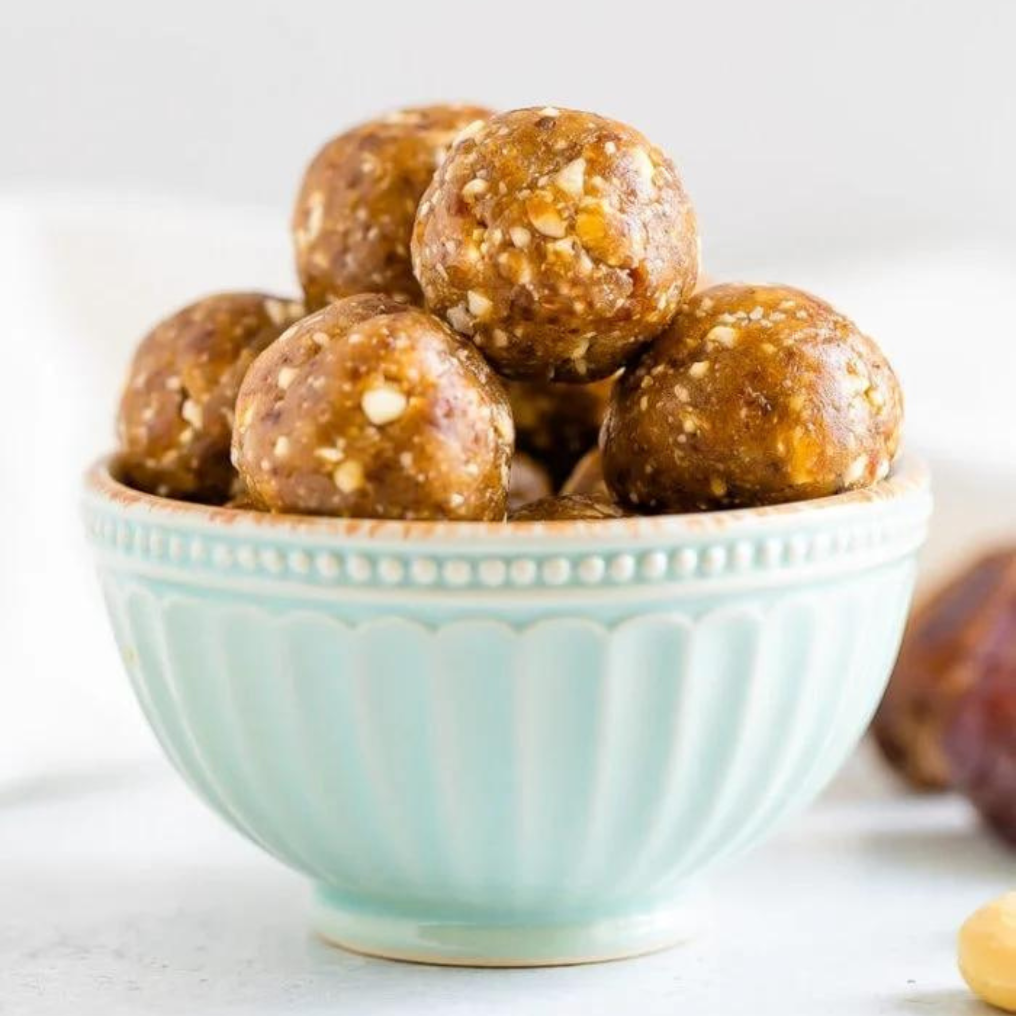 date balls piled into a sea green bowl on a white table with dates in the background