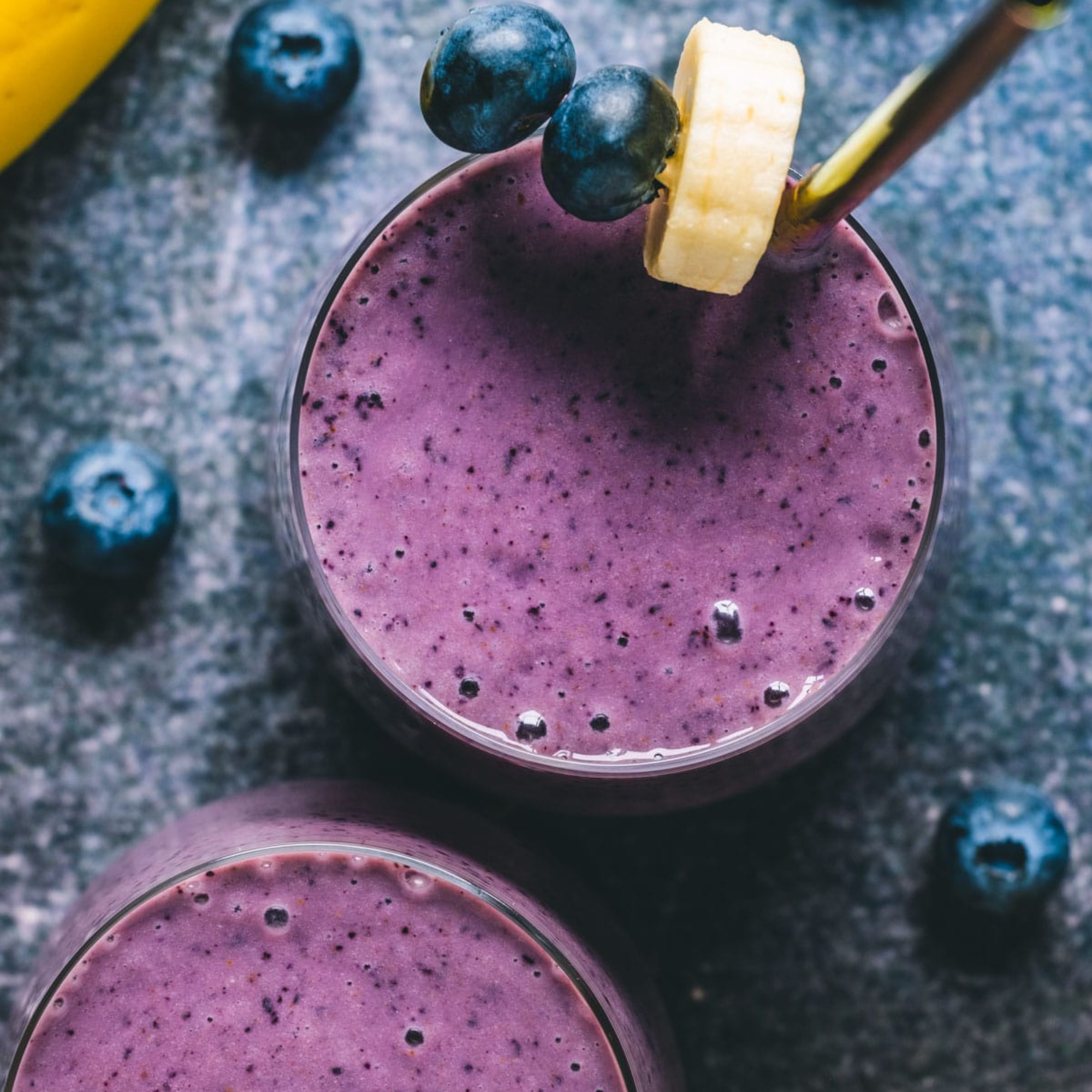a glass cup of blueberry banana smoothie with a spoon and a banana slice on a dark table top with scattered blueberries