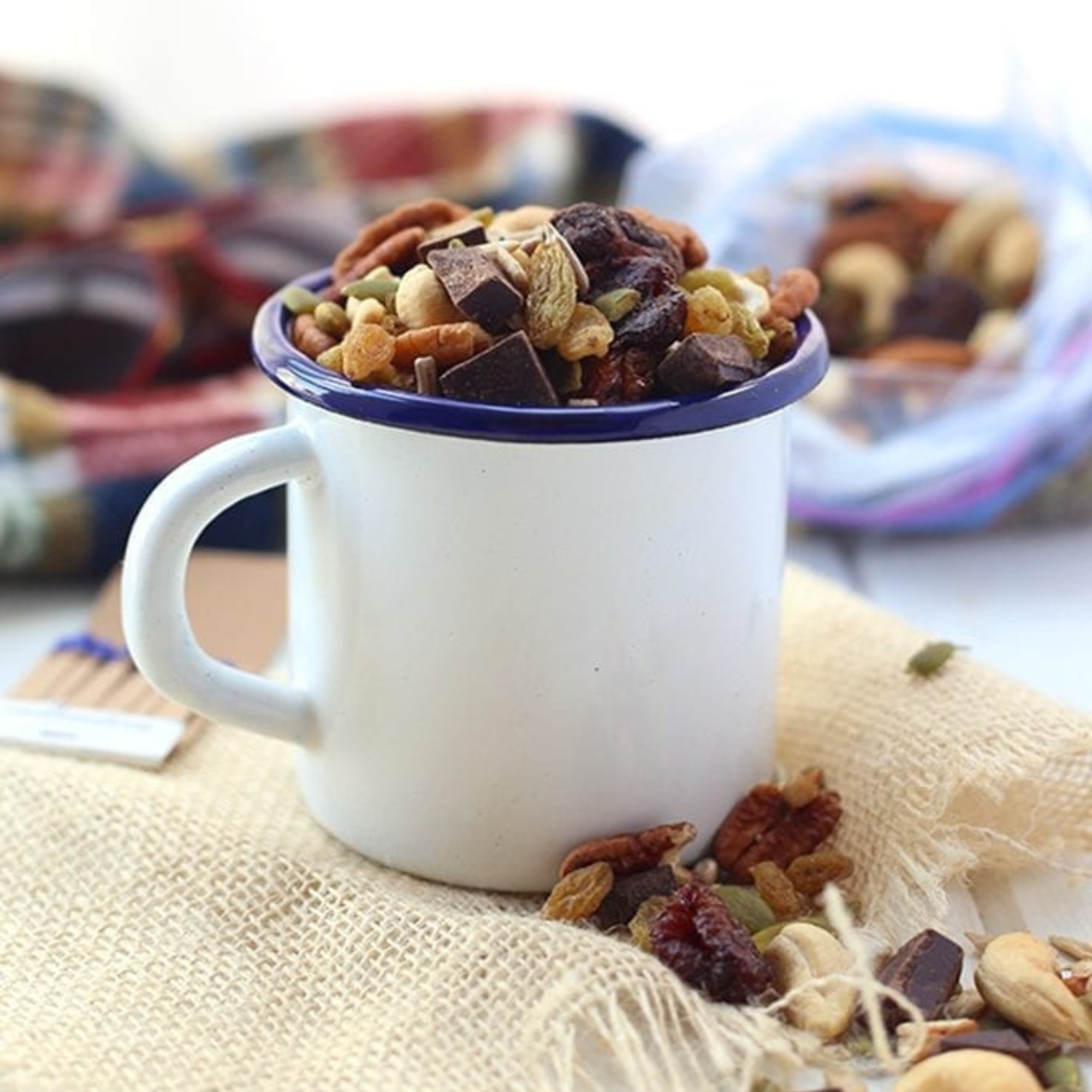 a white mug filled with healthy trail mix sitting on a cream colored kitchen cloth with trail mix scattered in front and fixing in the back