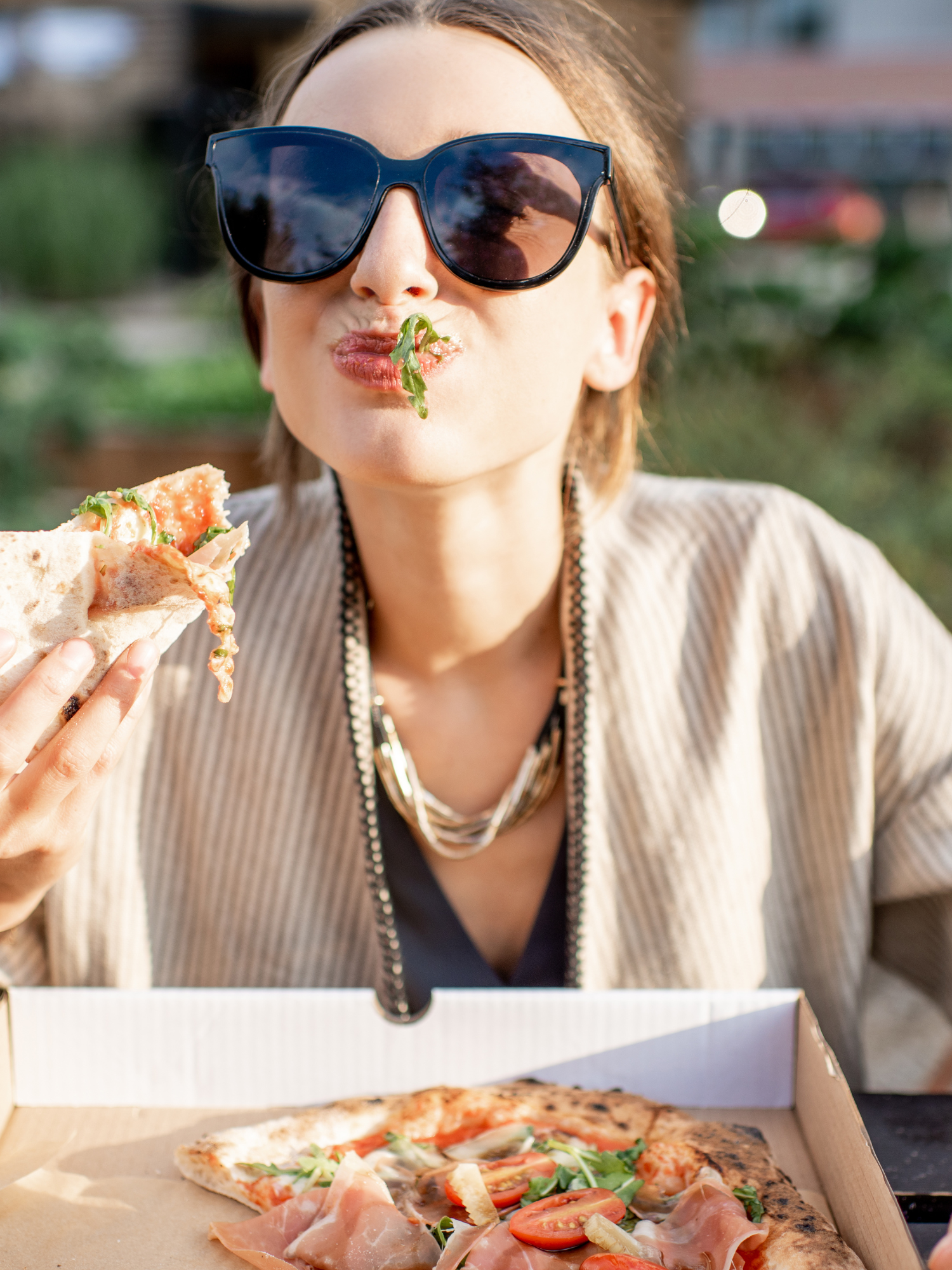 a woman sitting outside in big black sunglasses with an open pizza box in front of her and a slice in her hand while she is chewing with a piece of lettuce coming out of her mouth