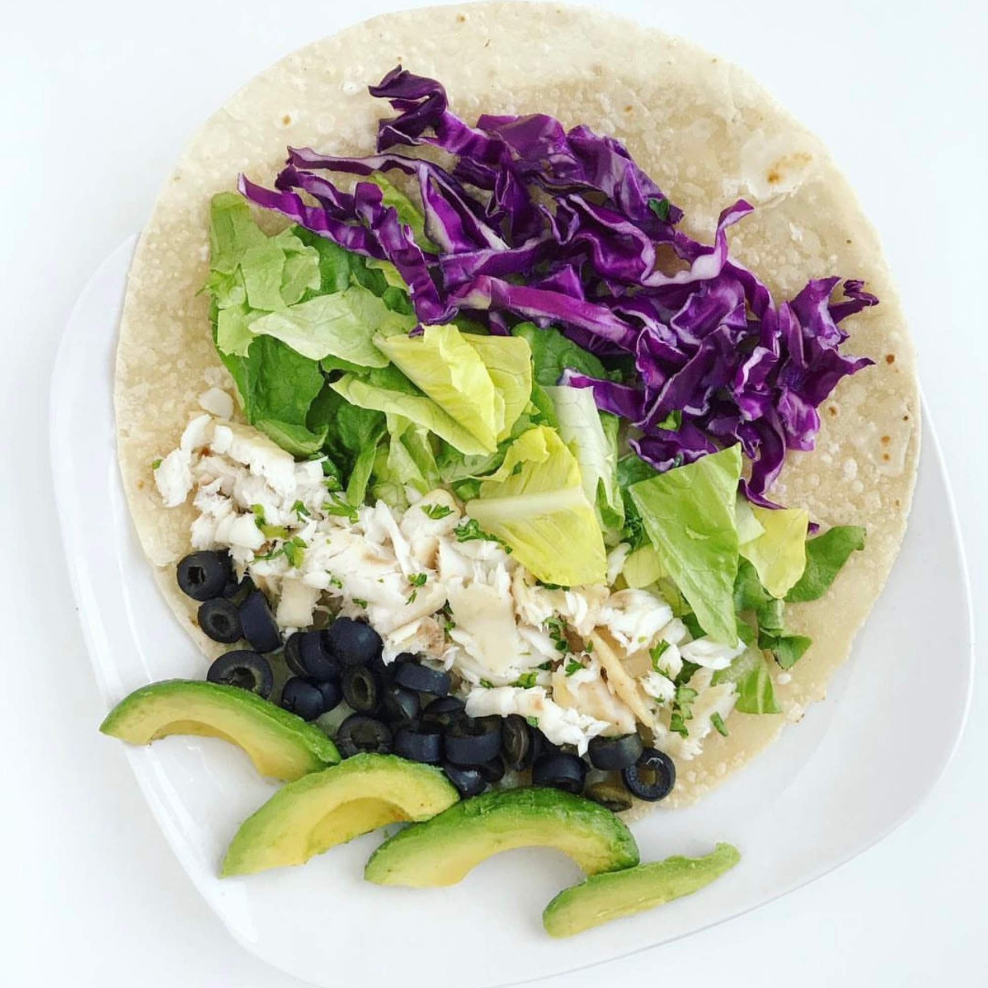a grain free tortilla on a white plate with a row of purple cabbage, lettuce, tilapia and black olives with some sliced avacado on the side