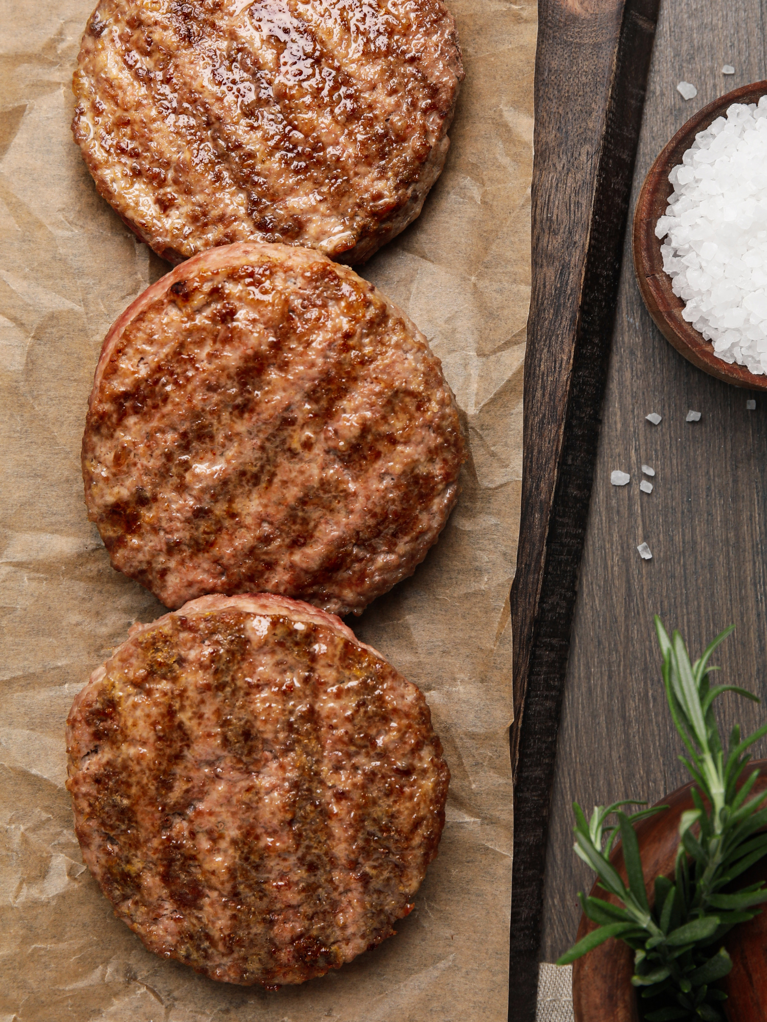 3 cooked and seasoned hamburger patties laying in a row on brown paper sitting next to some fresh herbs and a little pot of salt