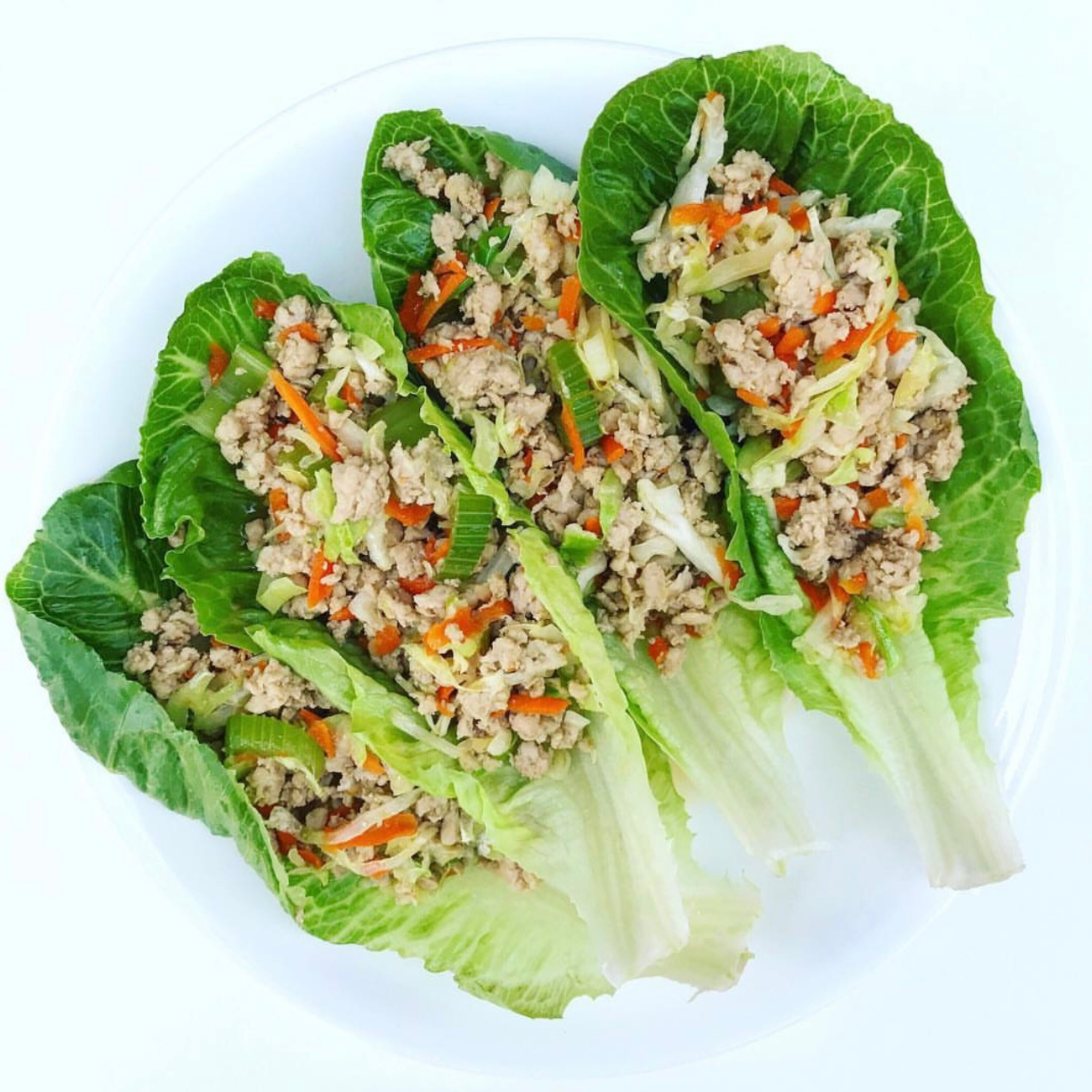 four lettuce wraps laid out on a white surface that are stuffed with a naked eggroll filling of cabbage, shredded carrots and chicken