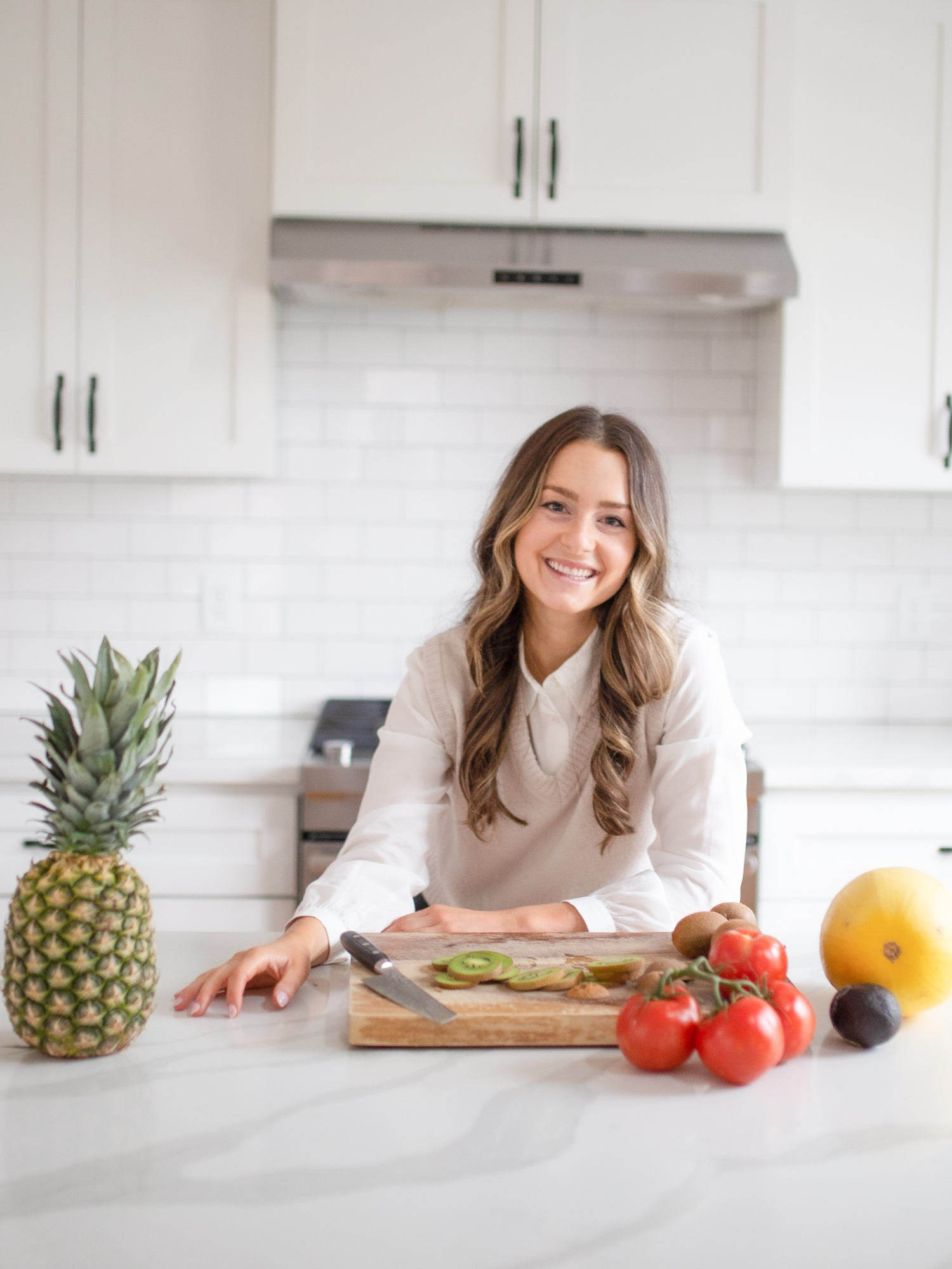 a woman dietician sitting at a kitchen counter with a cutting board and a pineapple, tomatoes, kiwis and a spaghetti squash