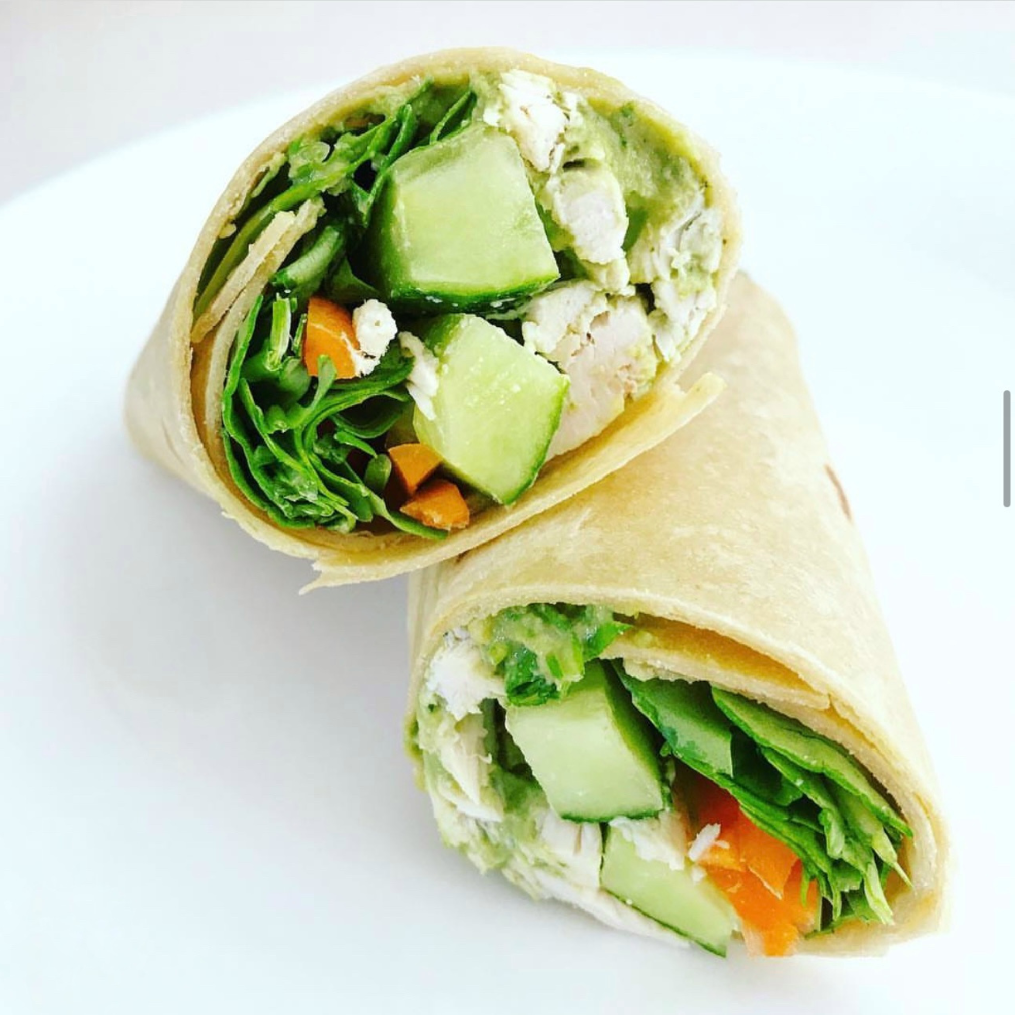 a gluten free tortilla filled with rotisserie chicken, homemade pesto, spinach, carrots and cucumbers cut in half and artfully placed on a white plate