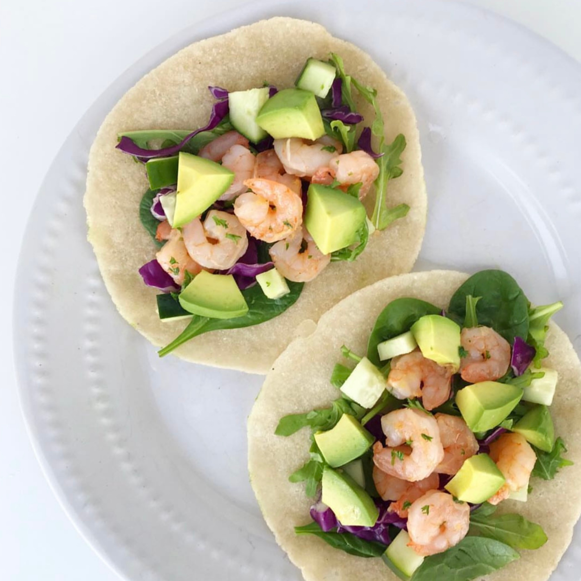 a white plate that has two gluten free tortillas topped with arugula, cucumbers, avocado and sauteed shrimp