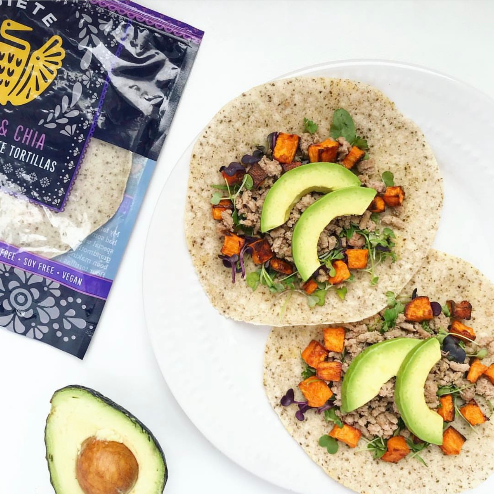 a white plate with two grain free tortilla open with roasted sweet potatoes, ground turkey, micro greens and sliced avacado. Next to the plate is the package of grain free tortillas and half an avocado