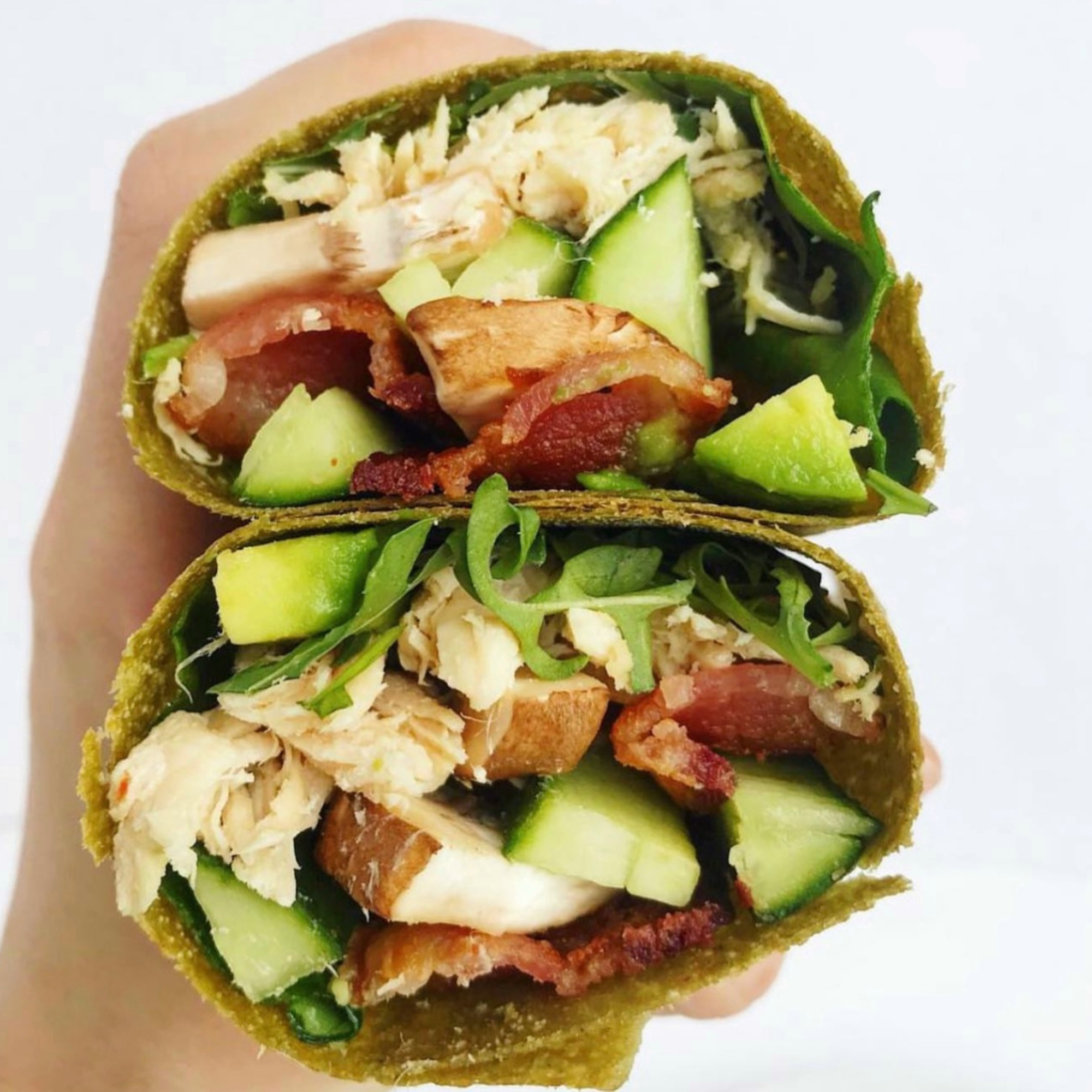 a wrap stuffed with filled it with arugala, spinach , turkey, mushrooms, avocado, cucumber and bacon cut in half and held in a womans hand