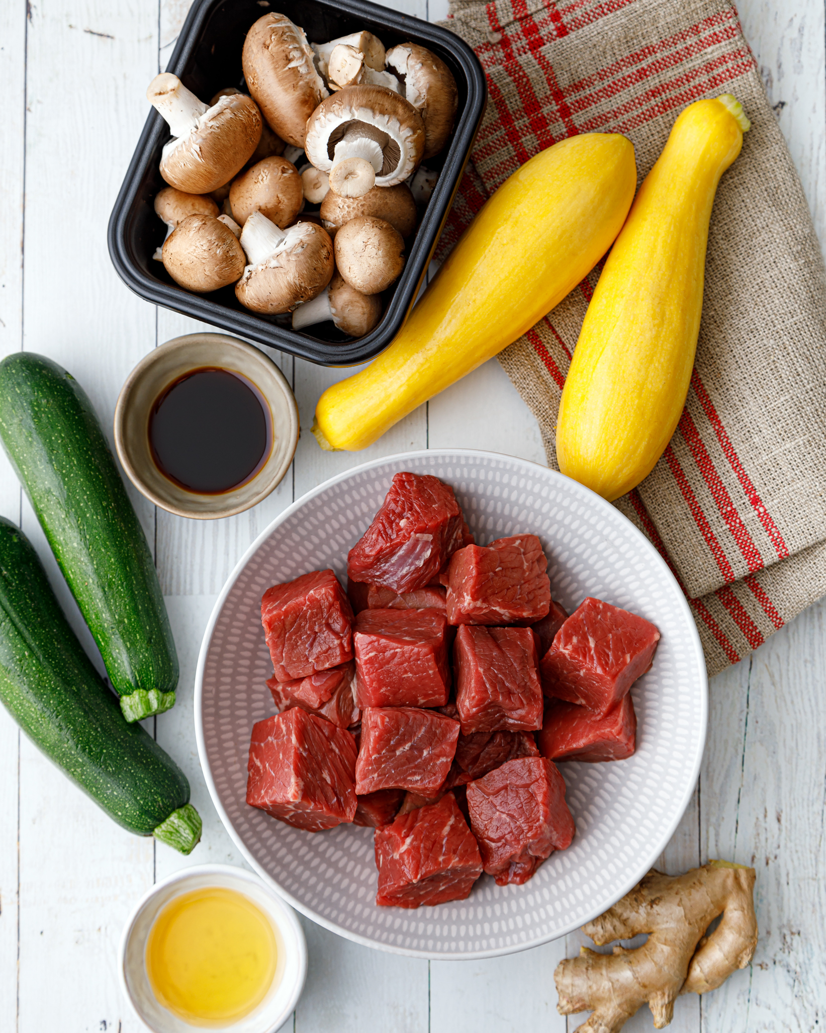 all the ingredients to make our beef kabobs, a white bowl filled with cubes of raw beef, two whole squash, two whole zucchini, a small bowl of our sweet and tangy sauce, and a carton of mushrooms