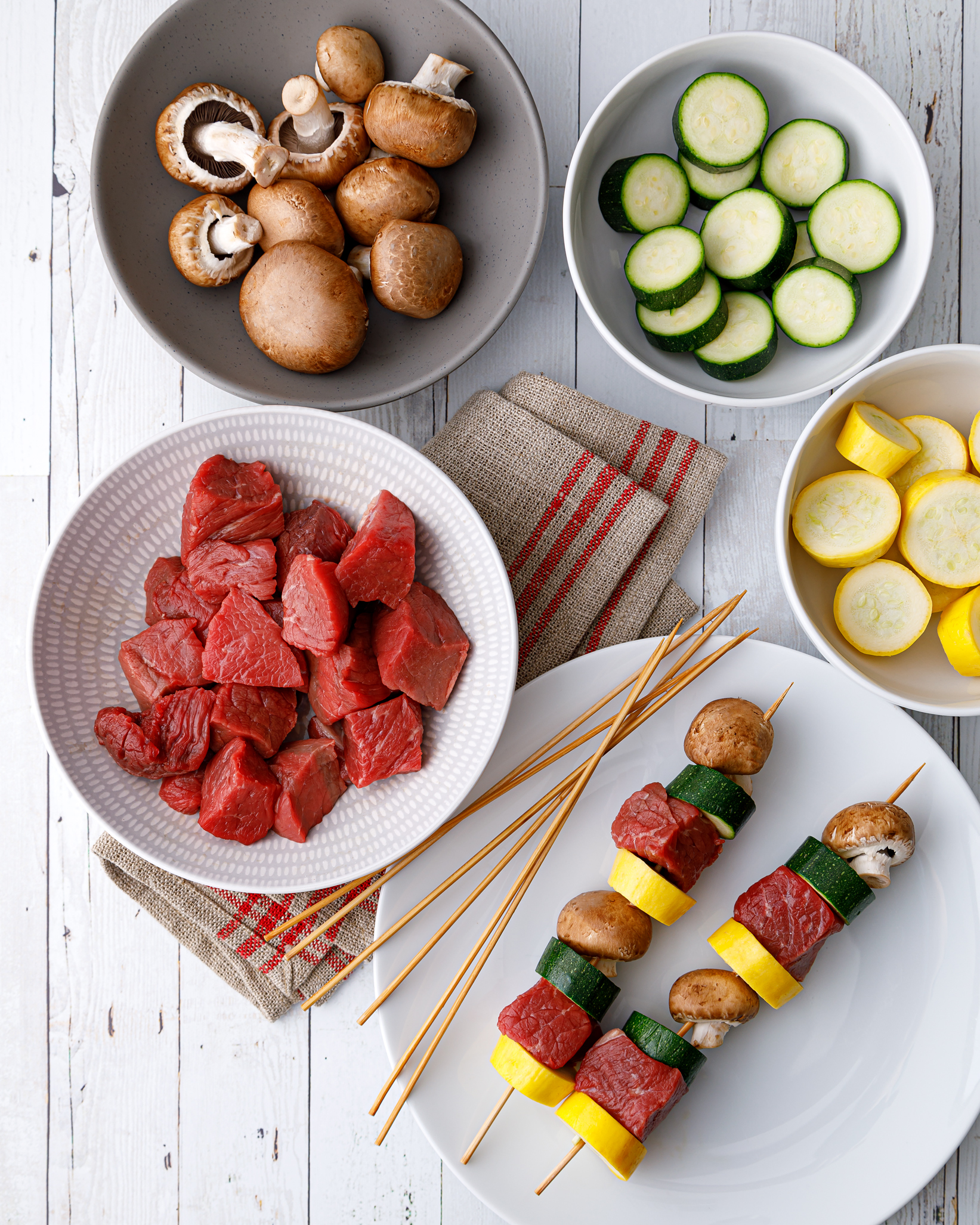 a plate of threaded beef and vegetable kebobs next to a small bowl of cubed beef, a small bowl of mushrooms, a small bowl of sliced zuchinni and a small bowl of sliced squash