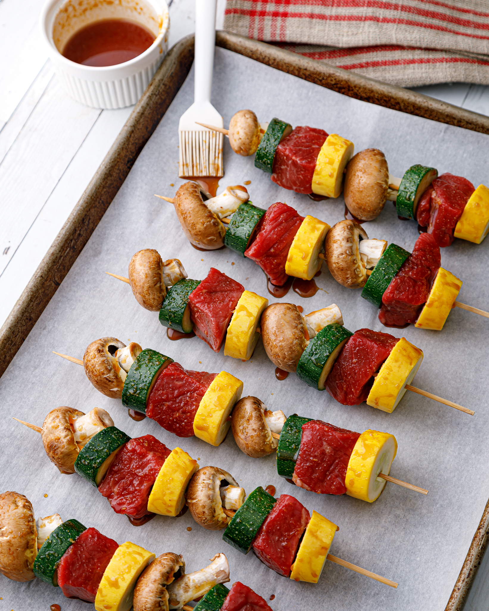 rows of beed kebobs on a parchment lined baking sheet with a small bowl of our honey ginger glaze and a wet glazing brush laying next to it