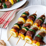 a whibeef kabobs that are skewered with squash and zucchini lined up neatly on a white plate with a red and white napkin and another similar plate in the background