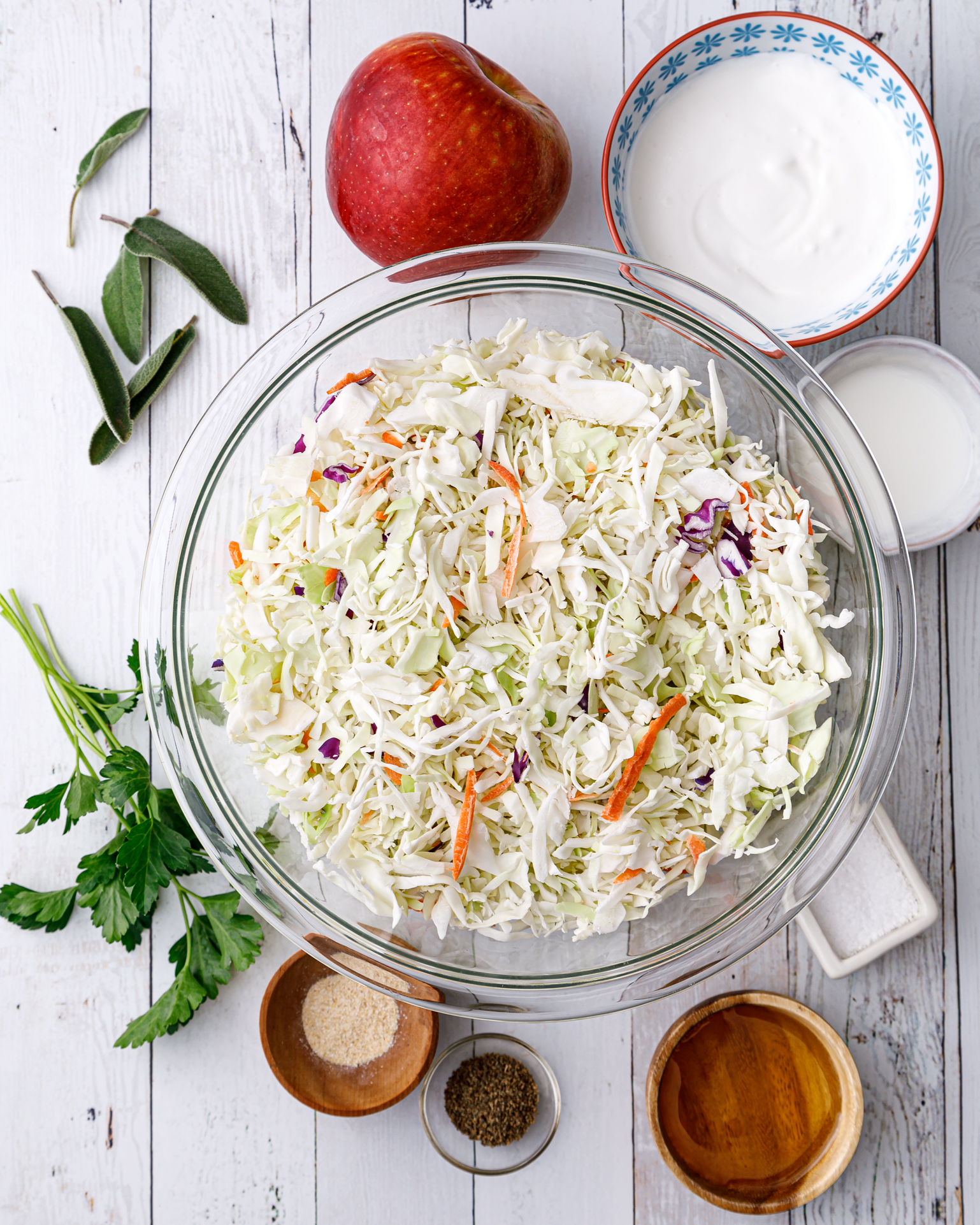 a bowl of shredded cabbage surrounded by a small bowl of plant based yogurt, a small bowl of plant based milk, an apple, fresh sage, and small bowls of various seasonings