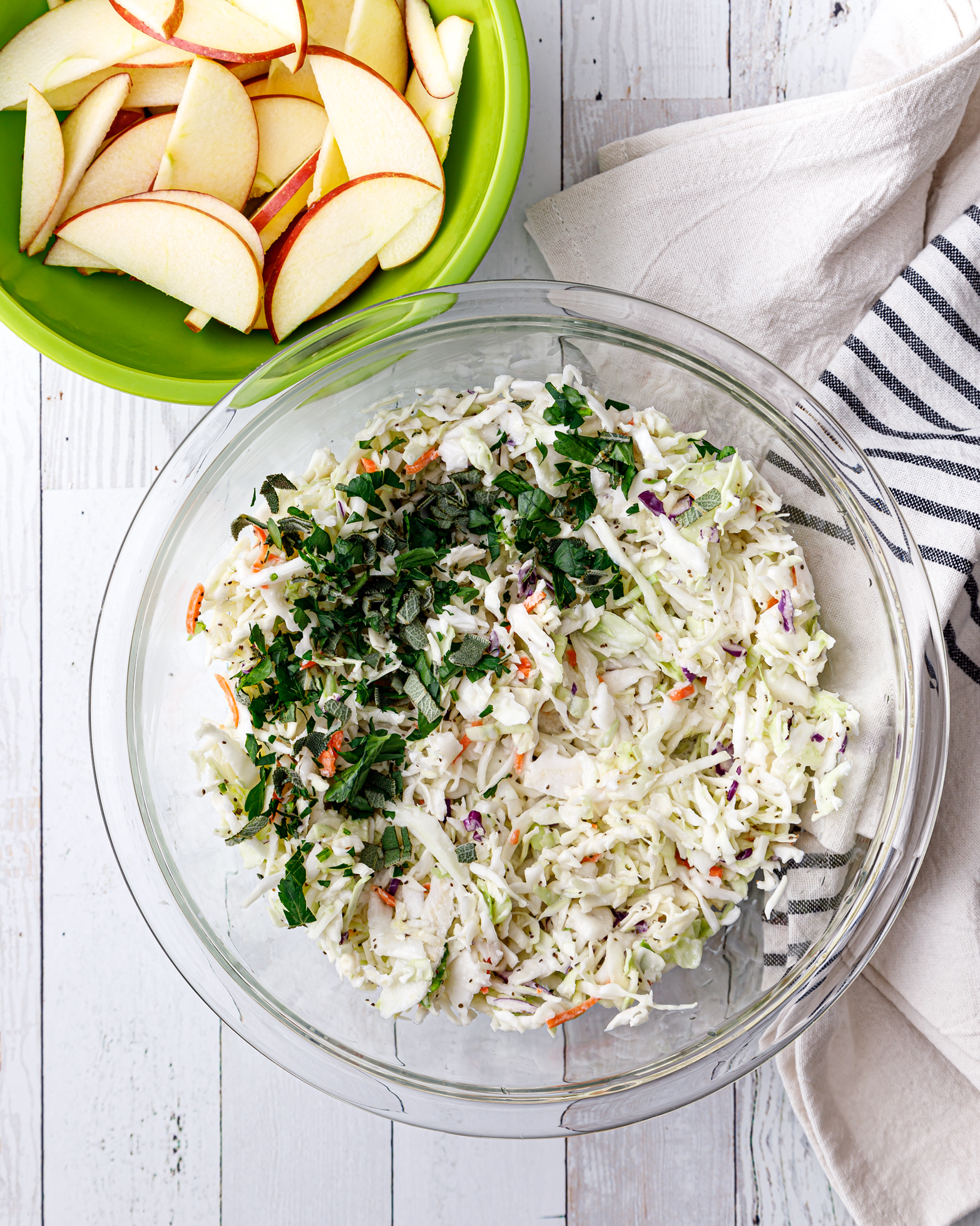 a glas bowl of creamy whole30 coleslaw next to a green bowl filled with apple sauces with a white a blue dish cloth next to both bowls