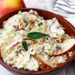 a brow wooden bowl full of creamy whole 30 coleslaw with apple slices on top and an apple sitting behind it