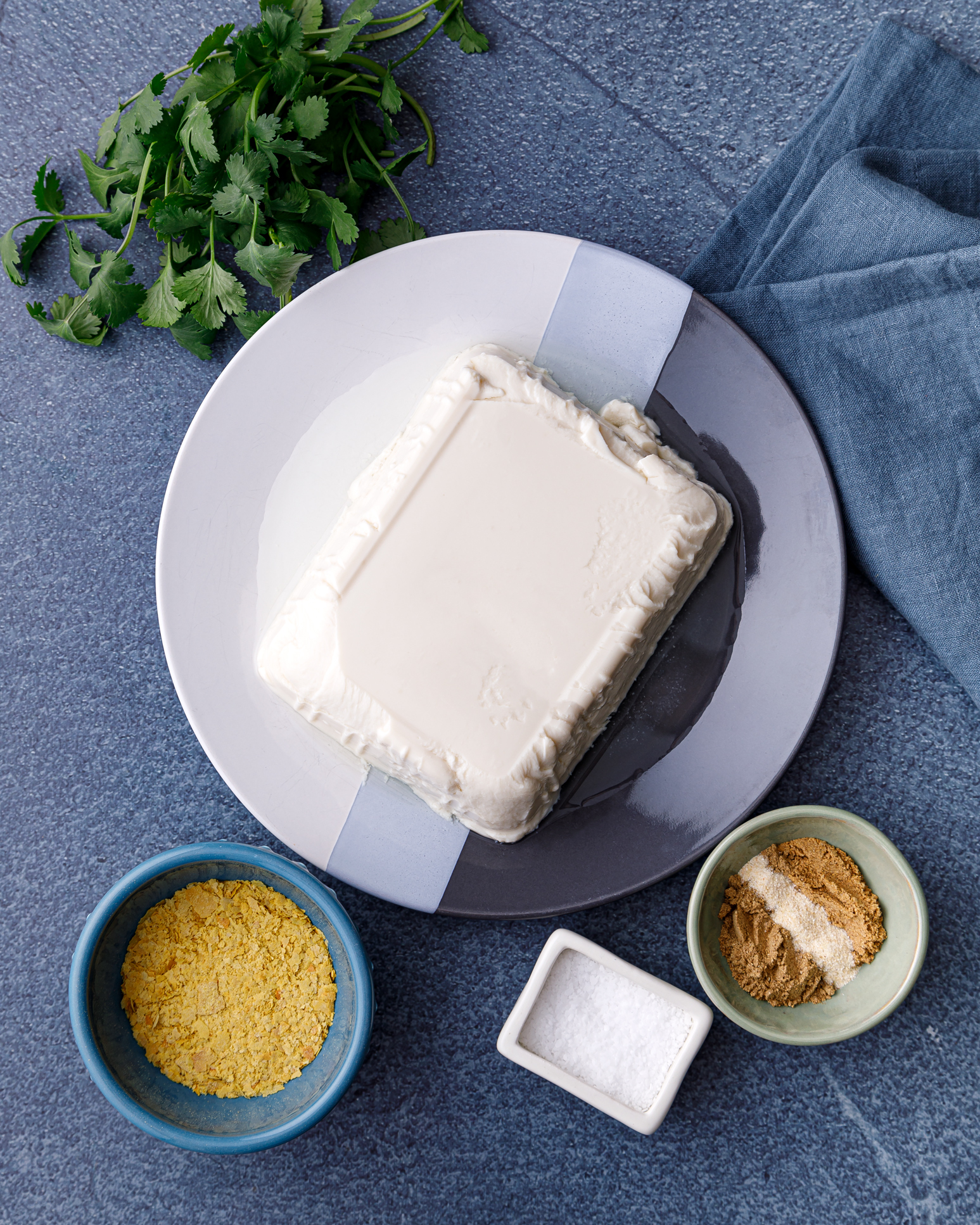 Ingredients for Tofu Queso laid out on a blue background. There is a plate wtih a block of queso, a small bowl of nutrtional yeast, a small container of salt, fresh cilantro and spices