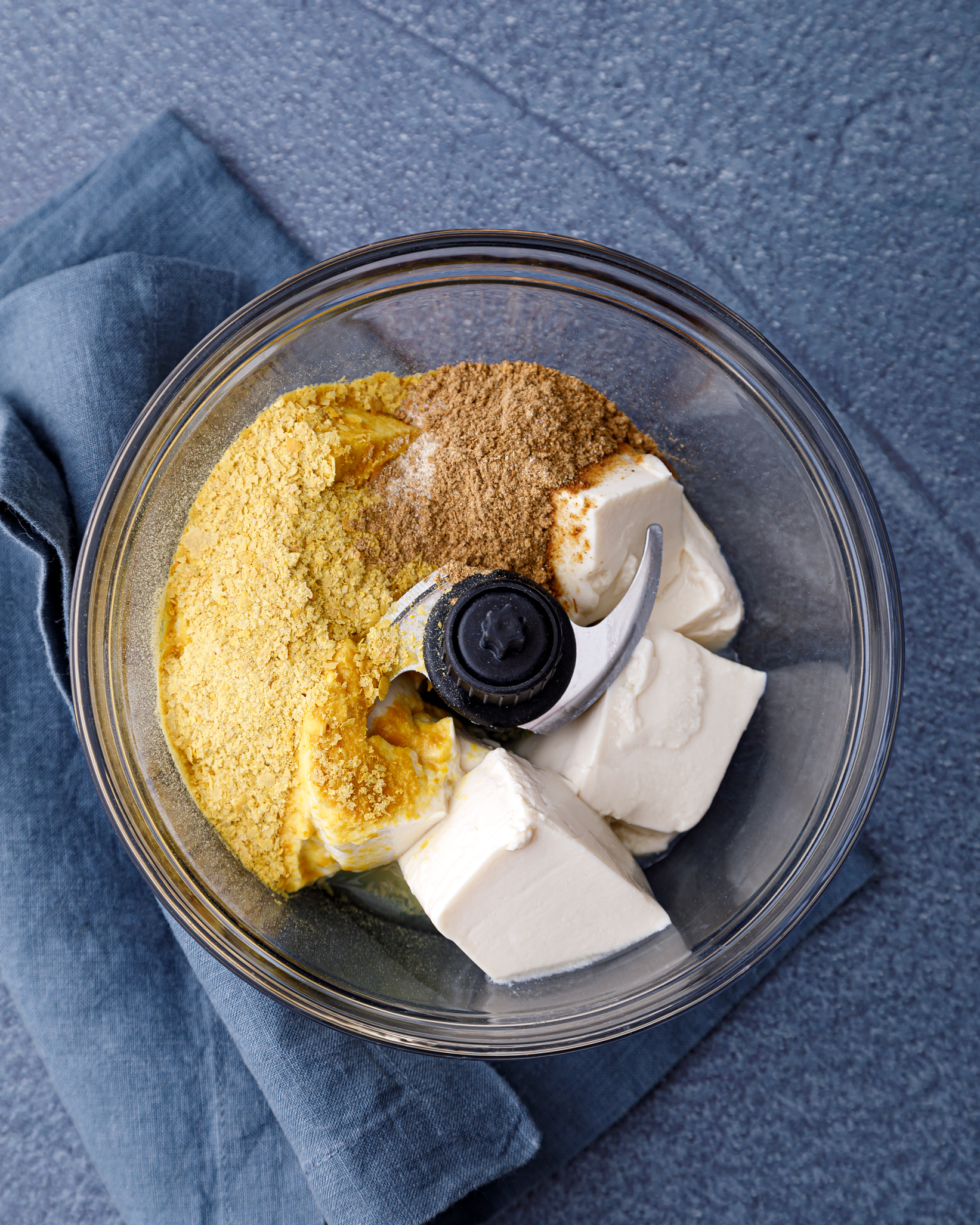 a food processor containing all the ingredients for. vegan, gluten free, nut free tofu queso. Tofu, nutritional yeast, cumin, coriander, onion powder and salt