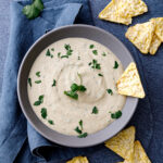 a gray bowl filled with rich and creamy Tofu Queso topped with pieces of cilantro and tortilla chips laid about
