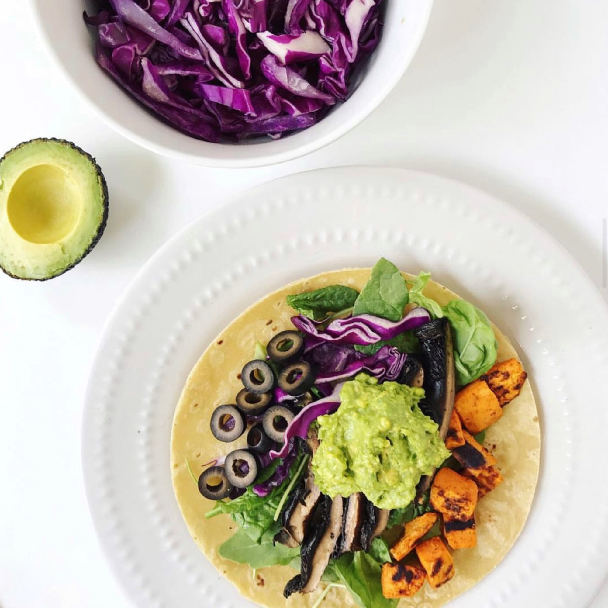 a white plate with a grain free tortilla that has baked sweet potatoes, portabello mushrooms and black olives with a dollop of guacamole on top and a white bowl of shredded purple cabbage behind it and half an avocado sitting beside it