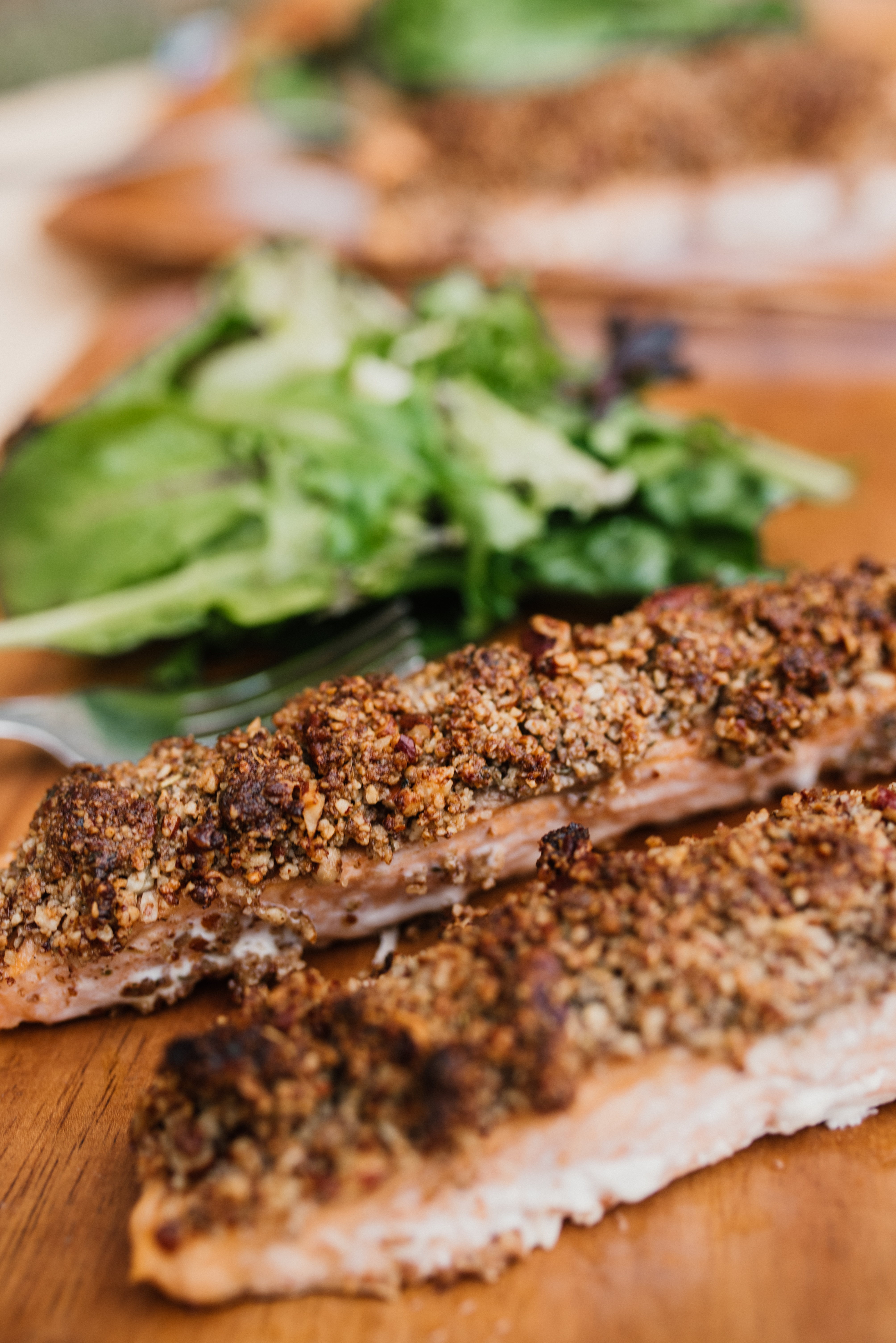 a wooden plate that has two pieces of honey pecan crusted salmon with a small side salad