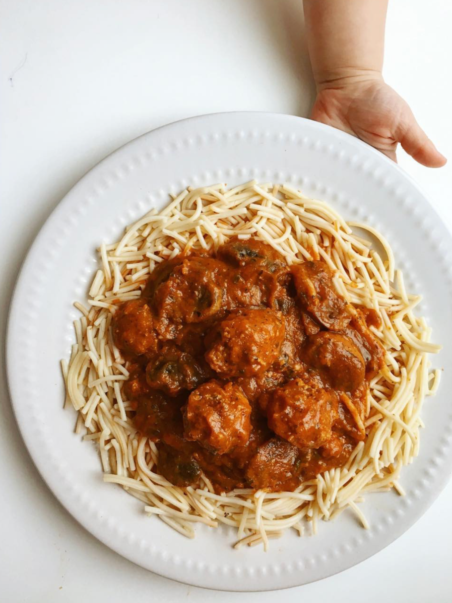 a plate of spaghetti with nomato sauce and meatballs with a childs hand holding the plate out