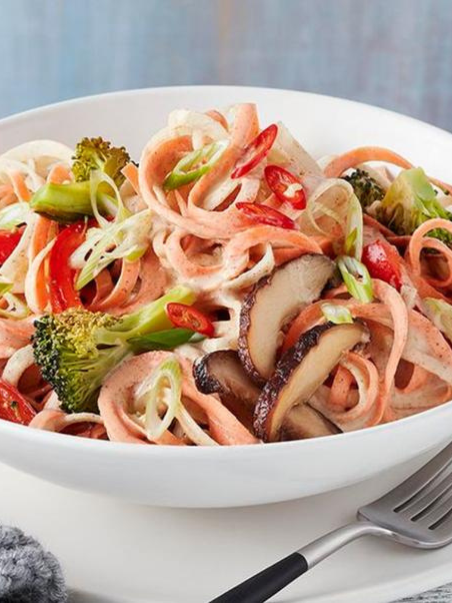 a big white bowl of Sesame Turnip Noodles with Broccoli & Mushrooms that is sitting on a white table with a fork next to it