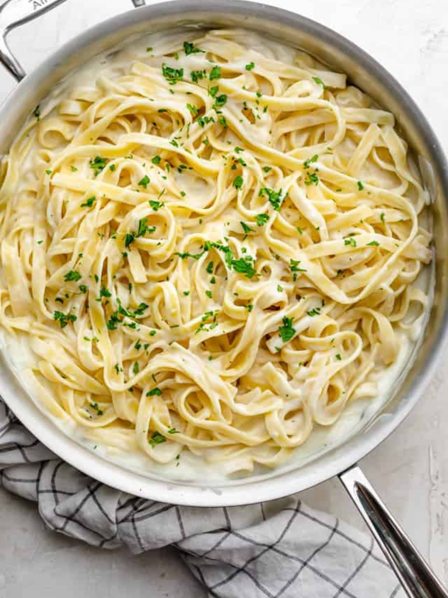 a stainless steel skillet full of cauliflower alfredo sitting on a white table with a checkered dish rag