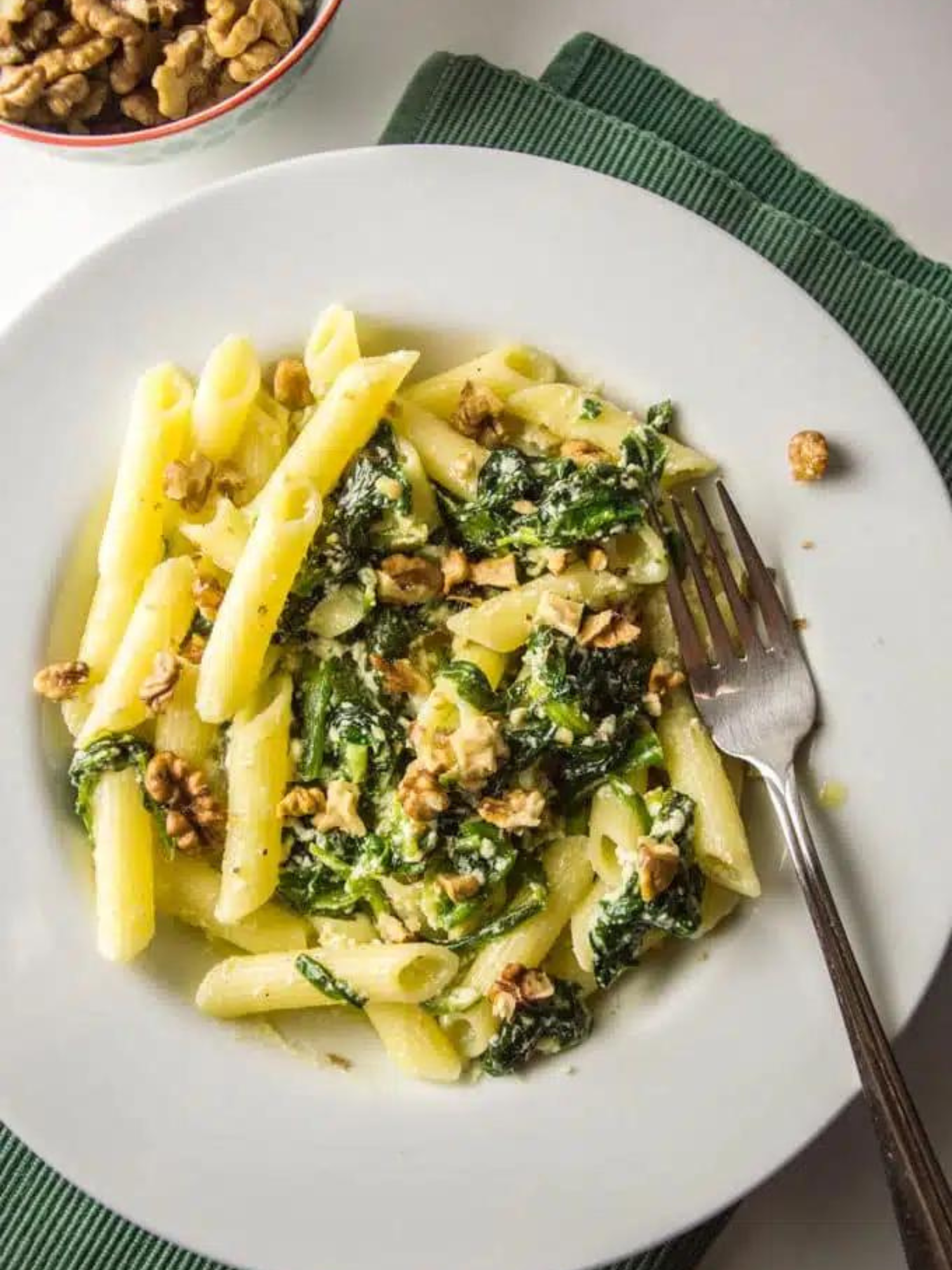a white bowl of Creamy Spinach and Ricotta Pasta with Walnuts, it's sitting on a white table on top of a dark green placemat with a small bowl of walnuts behind it