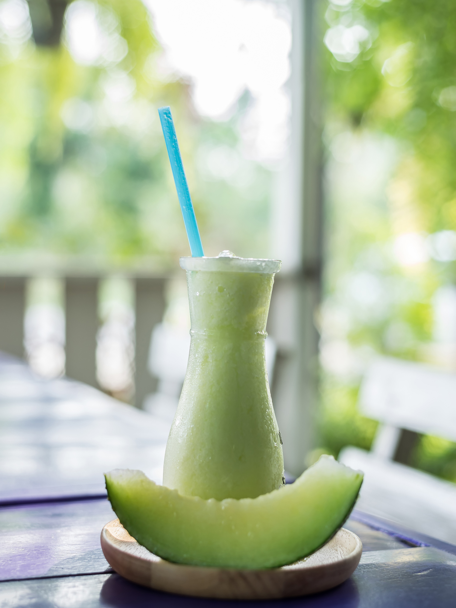 a honeydew alkaline smoothie in a glass cup with a blue straw on a sunny back porch with a slice of honey dew sitting in front of the smoothie