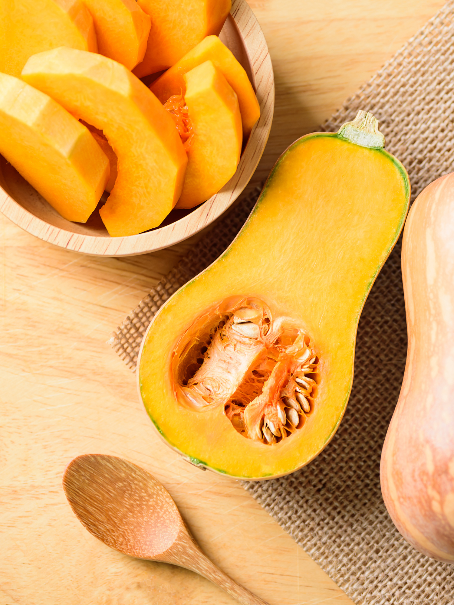 a butternut squash cut in half on a wooden table with a wooden spoon and a bowl of sliced butternut squash pieces