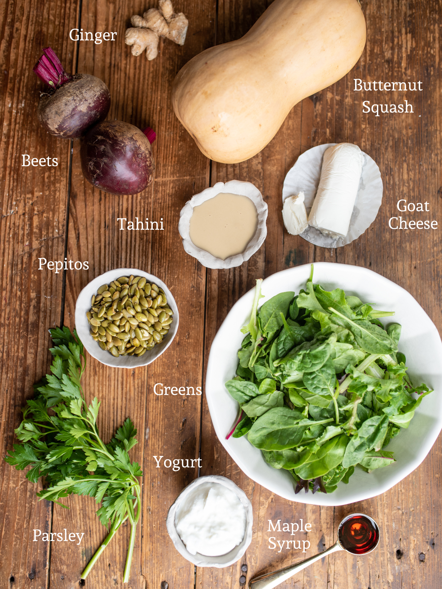 The ingredients for Beetroot and Butternut Salad laid out on a wooden table includeing a butternut squash, two beets, a piece of ginger, goat cheese, tahini, pepitos, mixed greens, parsley, yogurt, and. maple syrup