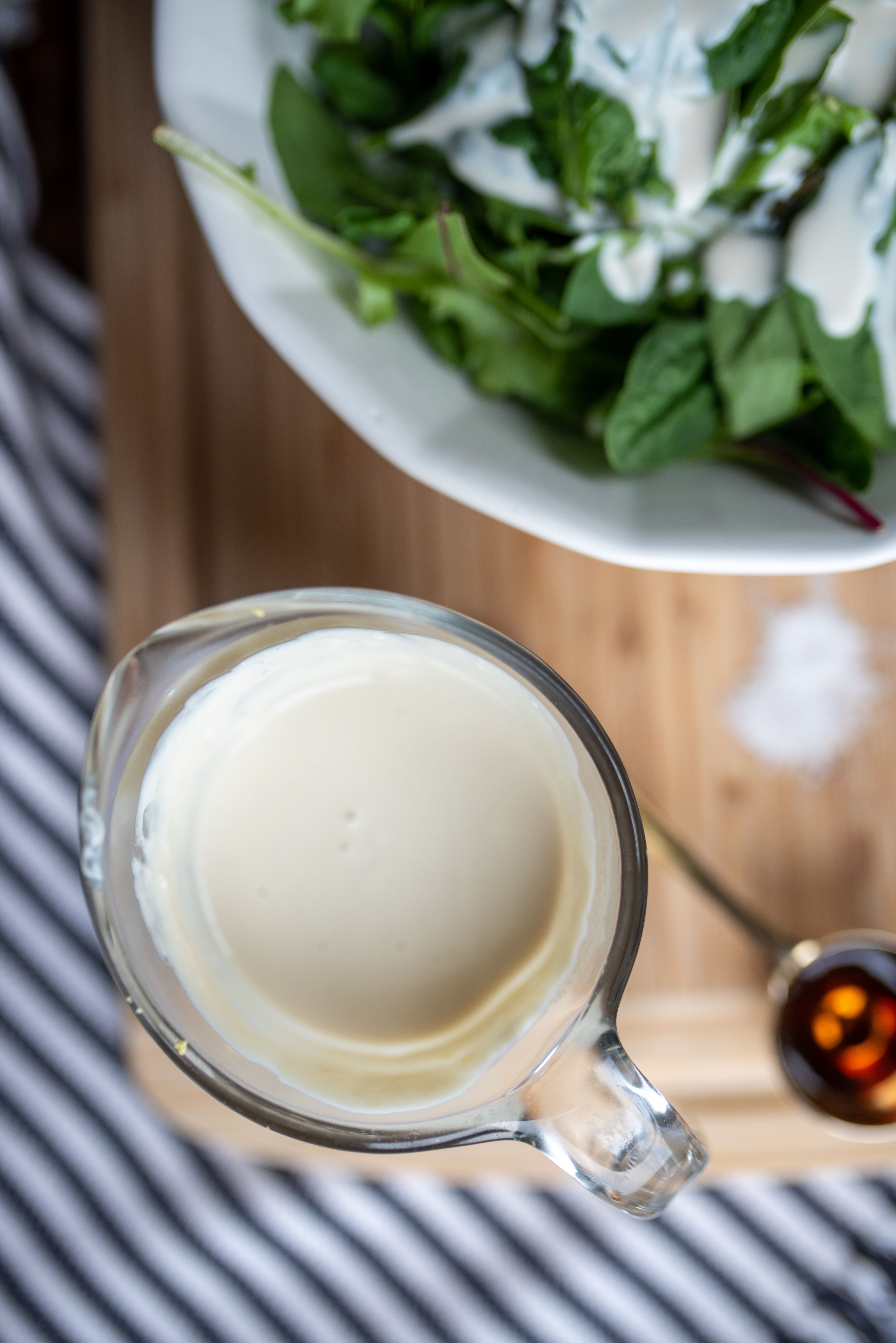 a glass measuring cup of low acid dressing on a cutting board with a salad drizzled in the dressing next to it and a blue and white striped cloth underneath