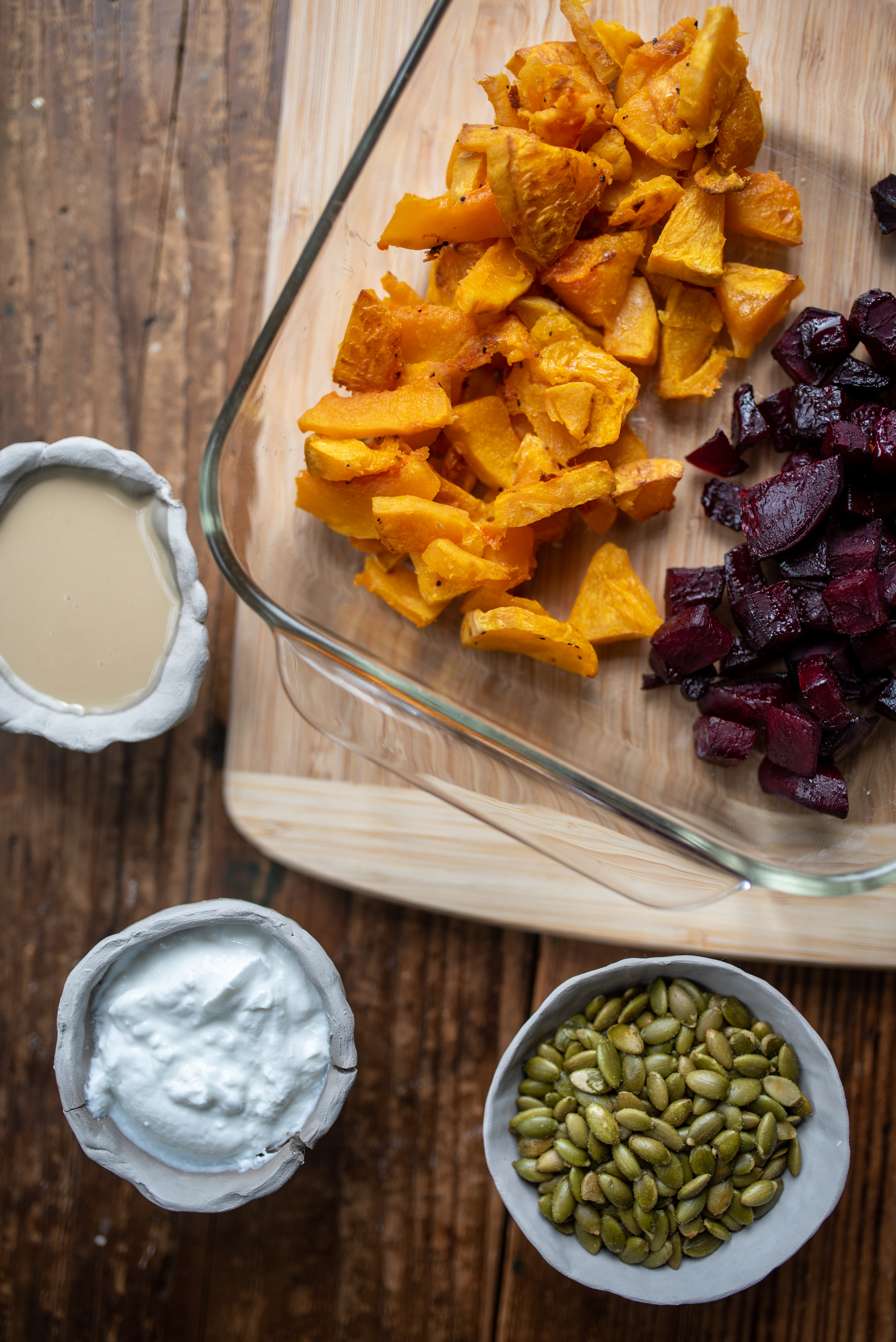 an 8x8 baking pan of roasted butternut squash and roasted beets on a cutting board with a small bowl or pepitos, a small bowl of yogurt and a small bowl of tahini next to it