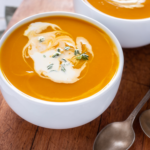 two white bowls filled with butternut squash soup and swired with coconut cream. They are sitting atop a cutting board with two silver spoons
