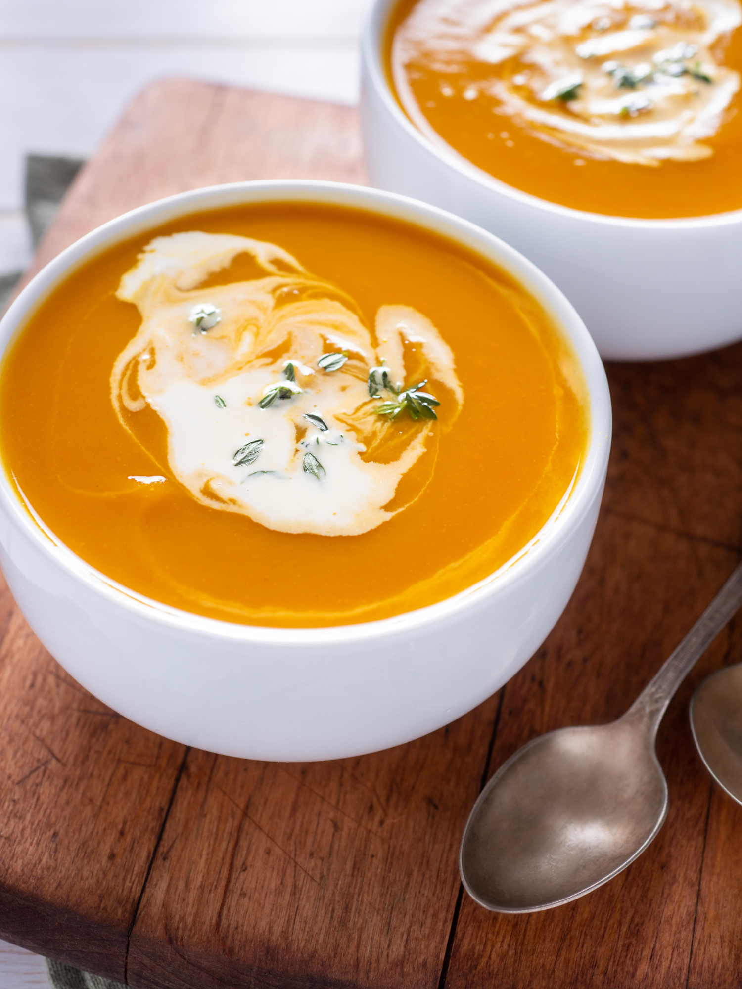 two white bowls filled with butternut squash soup and swired with coconut cream. They are sitting atop a cutting board with two silver spoons